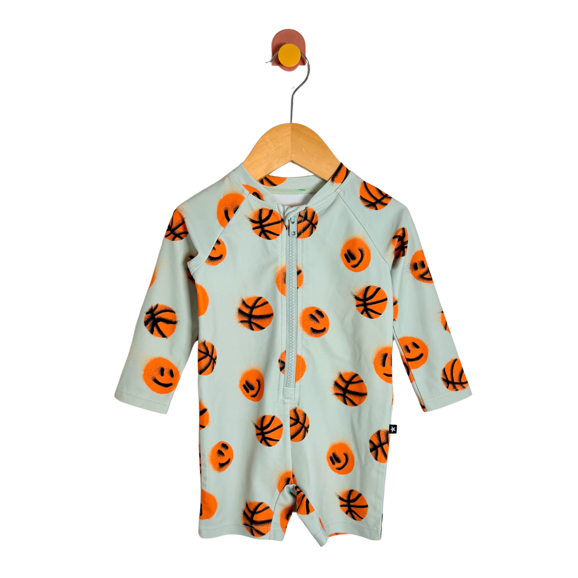 A baby long-sleeve zip-up romper in a pale mint green, decorated with an all-over orange basketball and smiley face pattern, hanging on a wooden hanger against a white background.