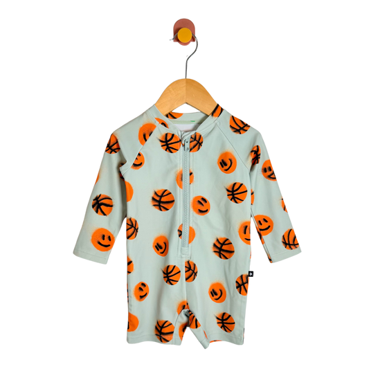 A baby long-sleeve zip-up romper in a pale mint green, decorated with an all-over orange basketball and smiley face pattern, hanging on a wooden hanger against a white background.