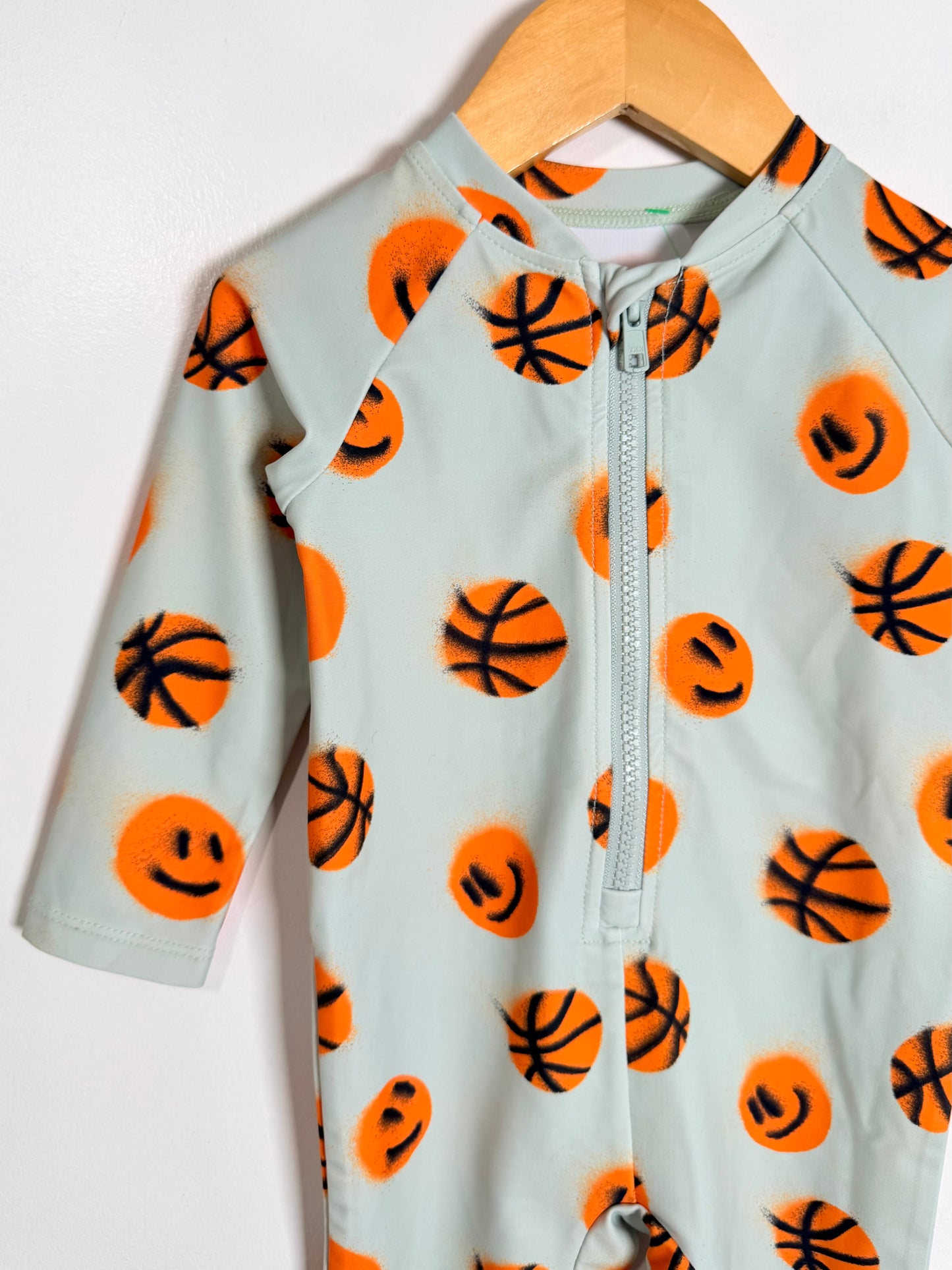 A baby long-sleeve zip-up romper in a pale mint green, decorated with a pattern of orange smiley-faced basketballs, hanging on a wooden hanger against a white wall.