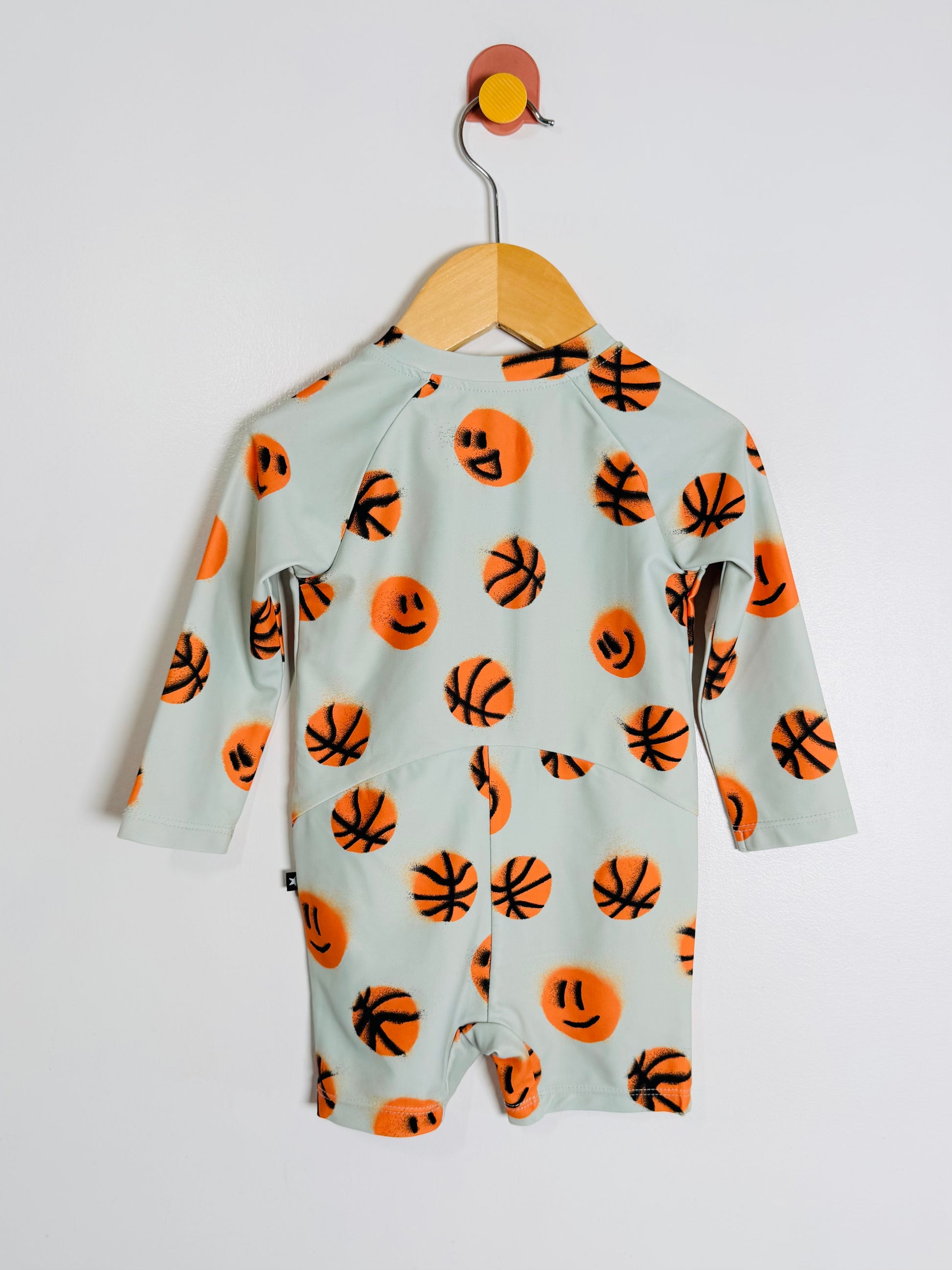 A baby long-sleeve romper in a pale sage green, featuring an all-over print of orange basketballs and smiley faces, hanging on a wooden hanger against a plain light gray wall.