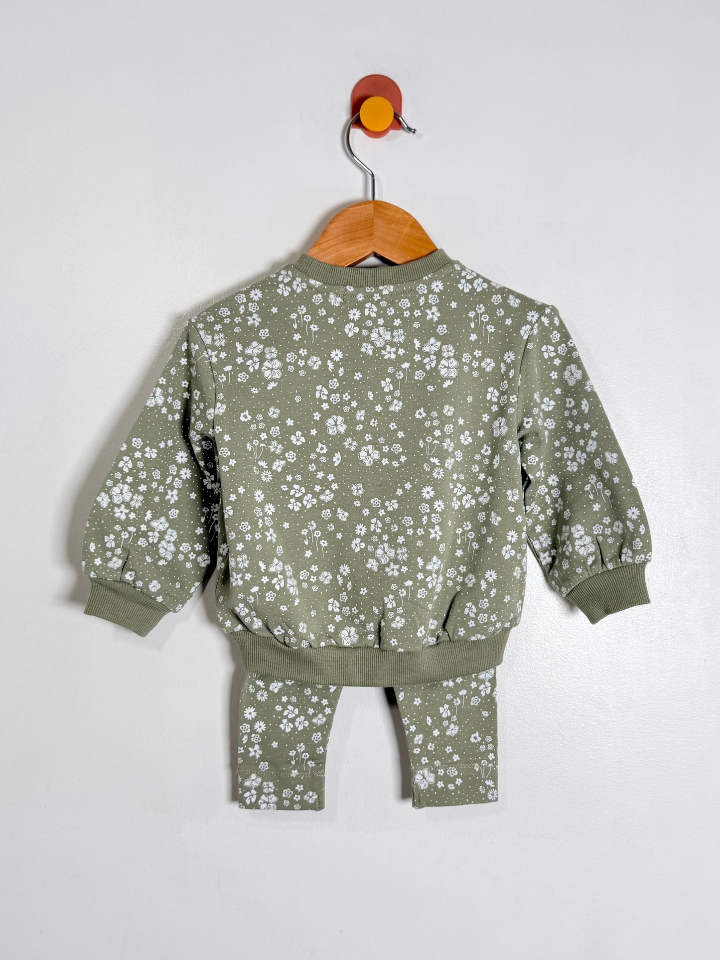A toddler two-piece matching set consisting of a long-sleeve sweatshirt and pants in sage green with a white floral print, hanging on a wooden hanger against a plain white wall.