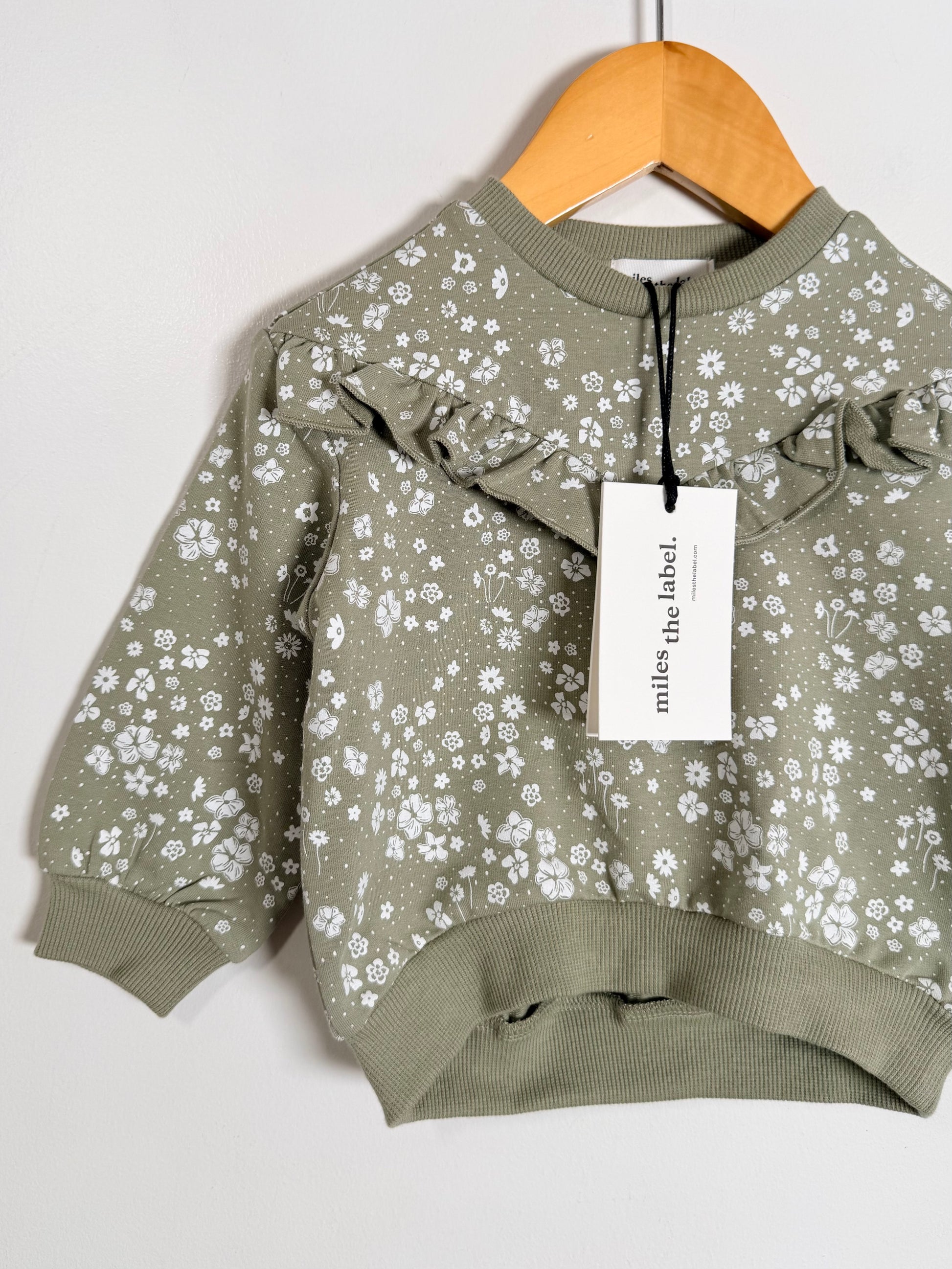 A toddler's long-sleeve sweatshirt in sage green with a white floral print, featuring ruffle details on the shoulders and ribbed cuffs and hem, hanging on a wooden hanger with a 'miles the label.' tag.