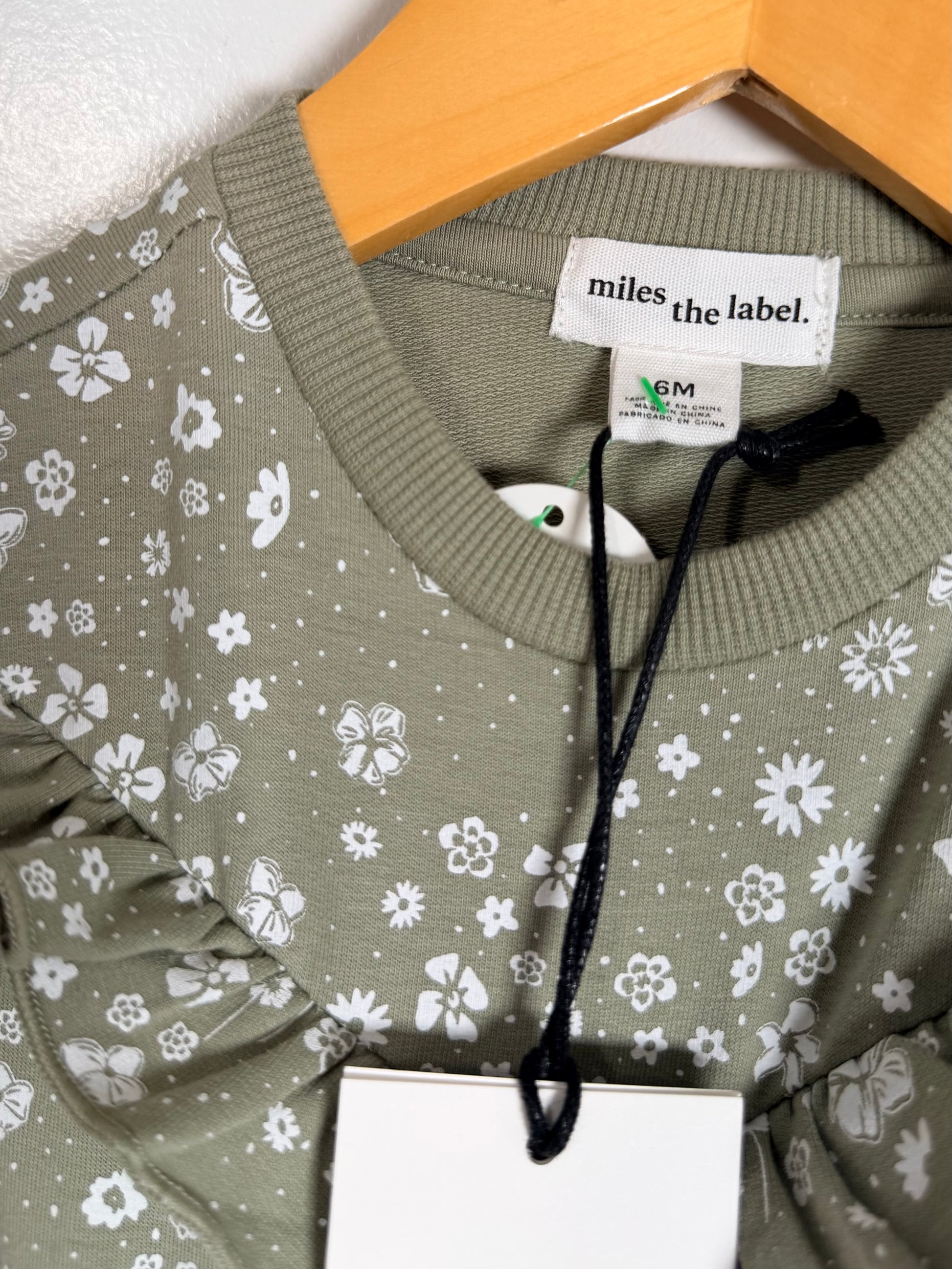 Close-up of a sage green long-sleeve top with a white floral print, hanging on a wooden hanger. The brand label 'miles the label.