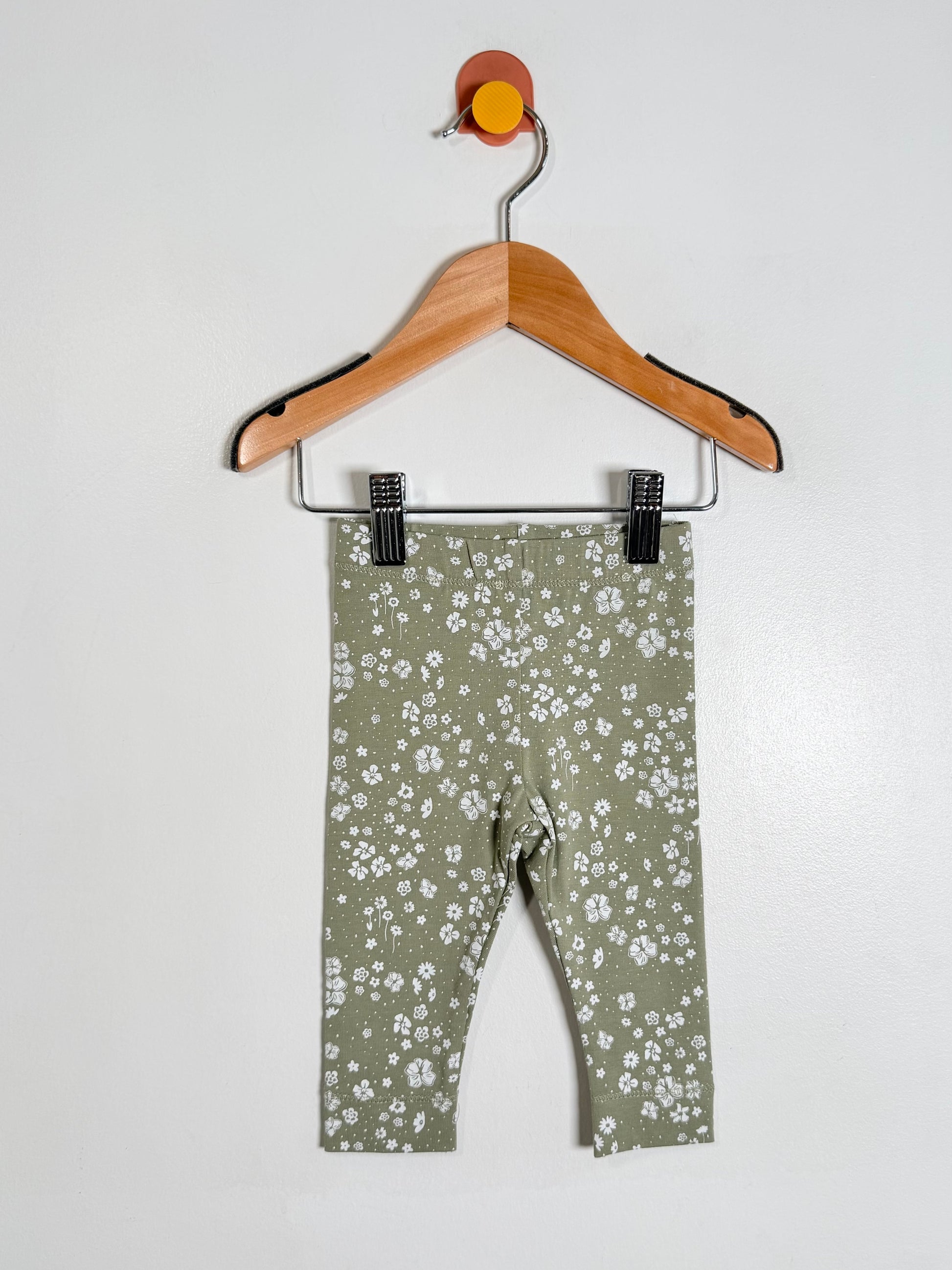 A pair of baby leggings in sage green with a white floral print, hanging from a wooden hanger against a plain white wall.