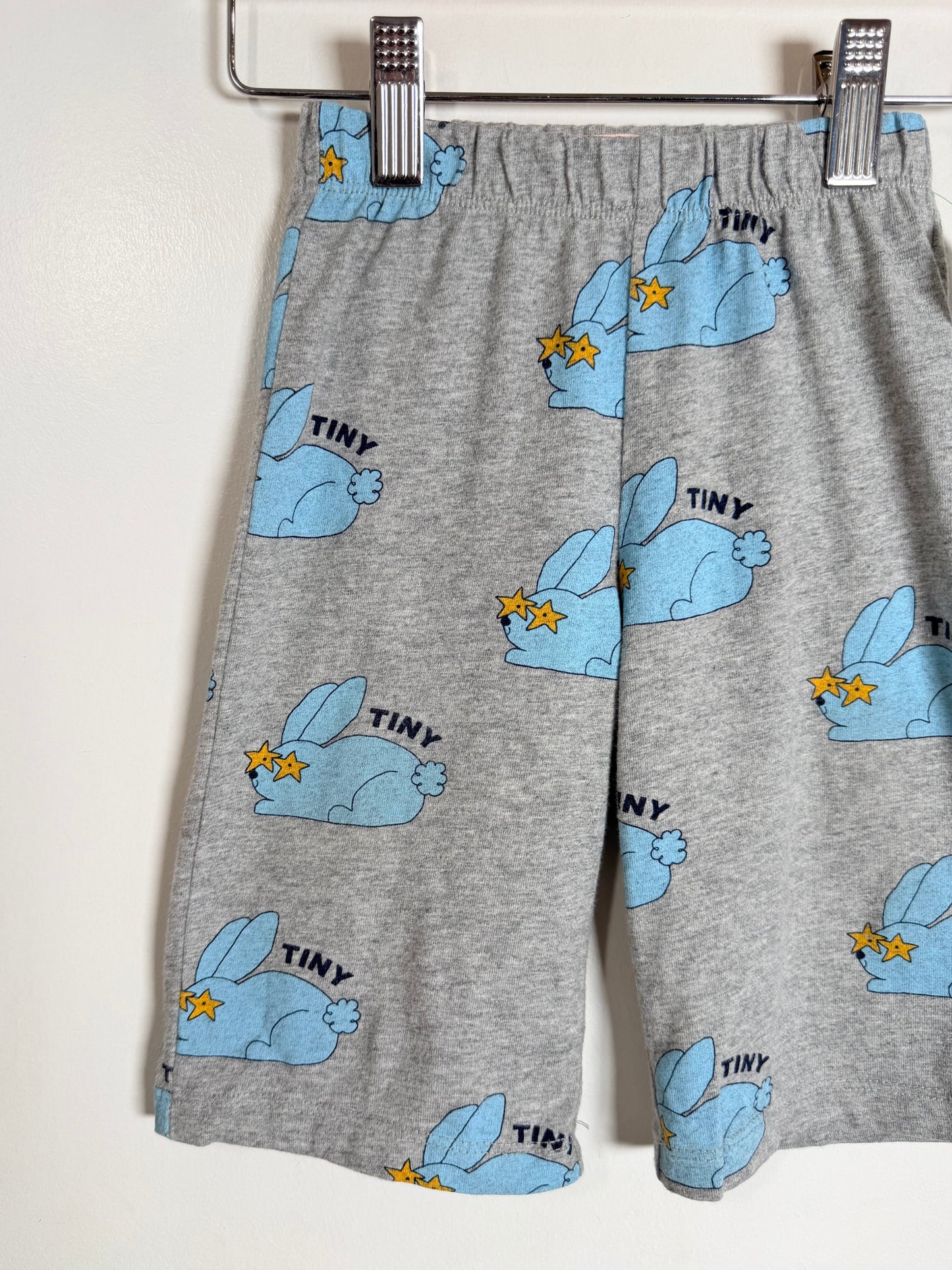 Gray heathered shorts with a repeating cartoon rabbit print in light blue, each wearing sunglasses and the word 'TINY' above it, hanging from a metal hanger against a plain light wall.