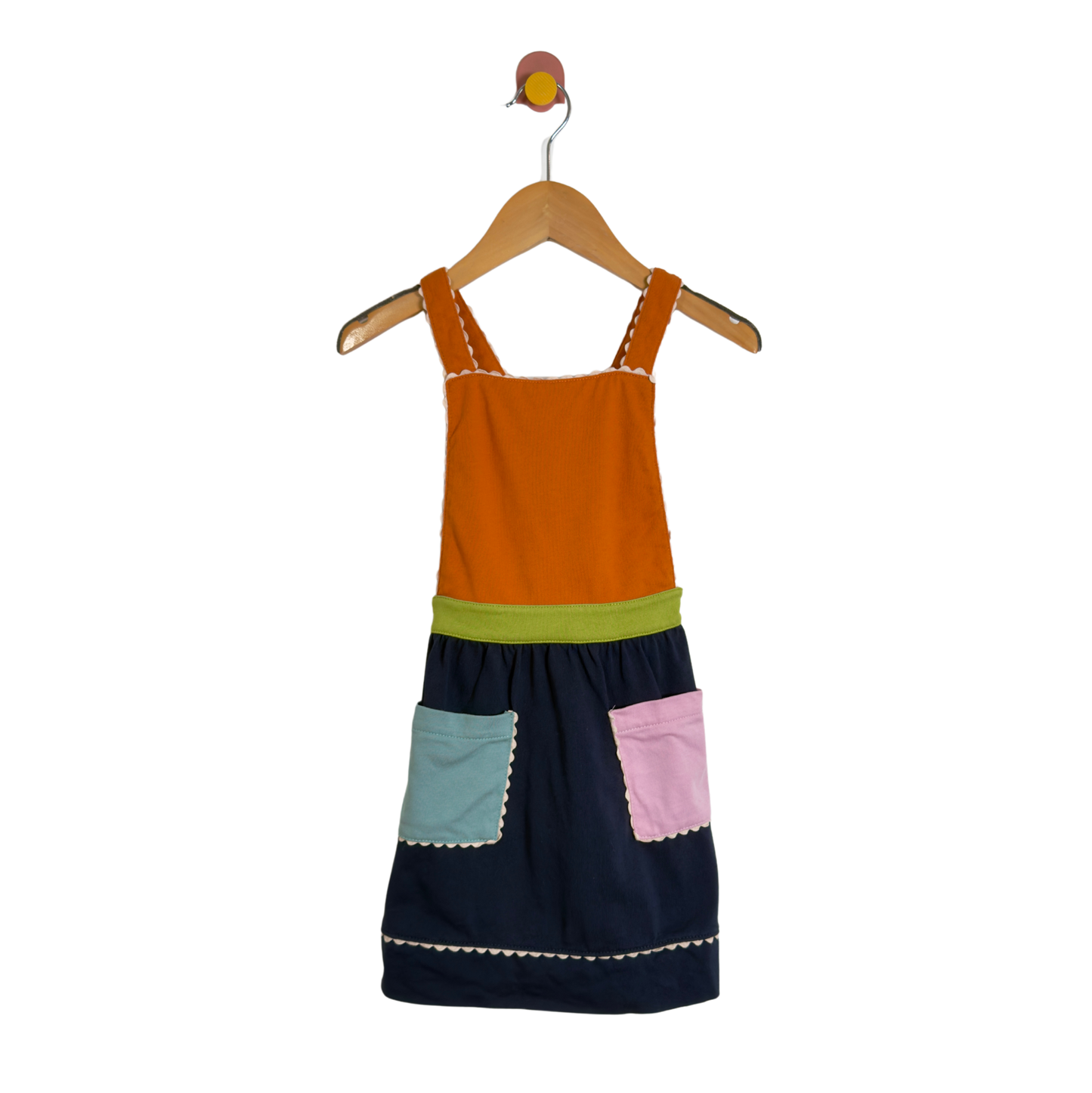 A children's patchwork-style pinafore dress hanging on a wooden hanger against a plain white background. The dress has an orange yoke, a lime green waistband, a dark navy skirt, a light blue front pocket, and a pink front pocket, all with white contrast stitching.