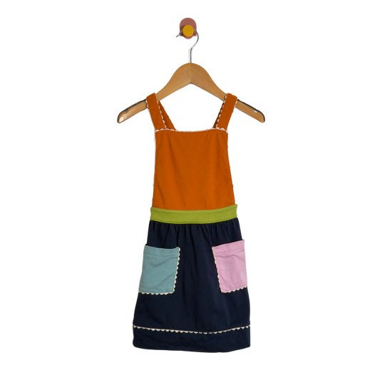 A children's patchwork-style pinafore dress hanging on a wooden hanger against a plain white background. The dress has an orange yoke, a lime green waistband, a dark navy skirt, a light blue front pocket, and a pink front pocket, all with white contrast stitching.