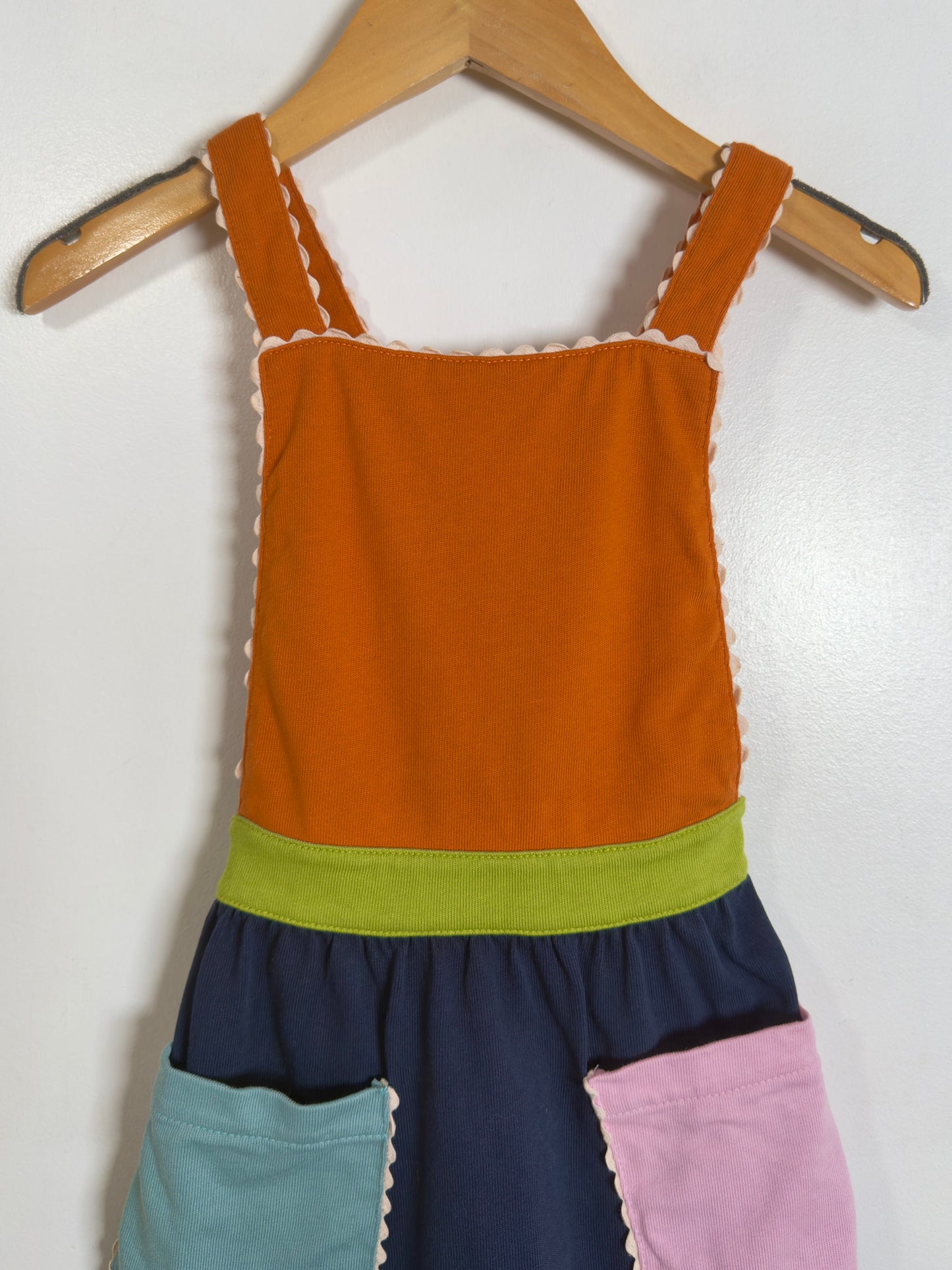 A sleeveless children's dress or pinafore-style top hanging on a wooden hanger against a plain white wall. The garment has an orange upper section, a lime green waistband, a navy lower section, and two front pockets in light blue and pink with scalloped edges.
