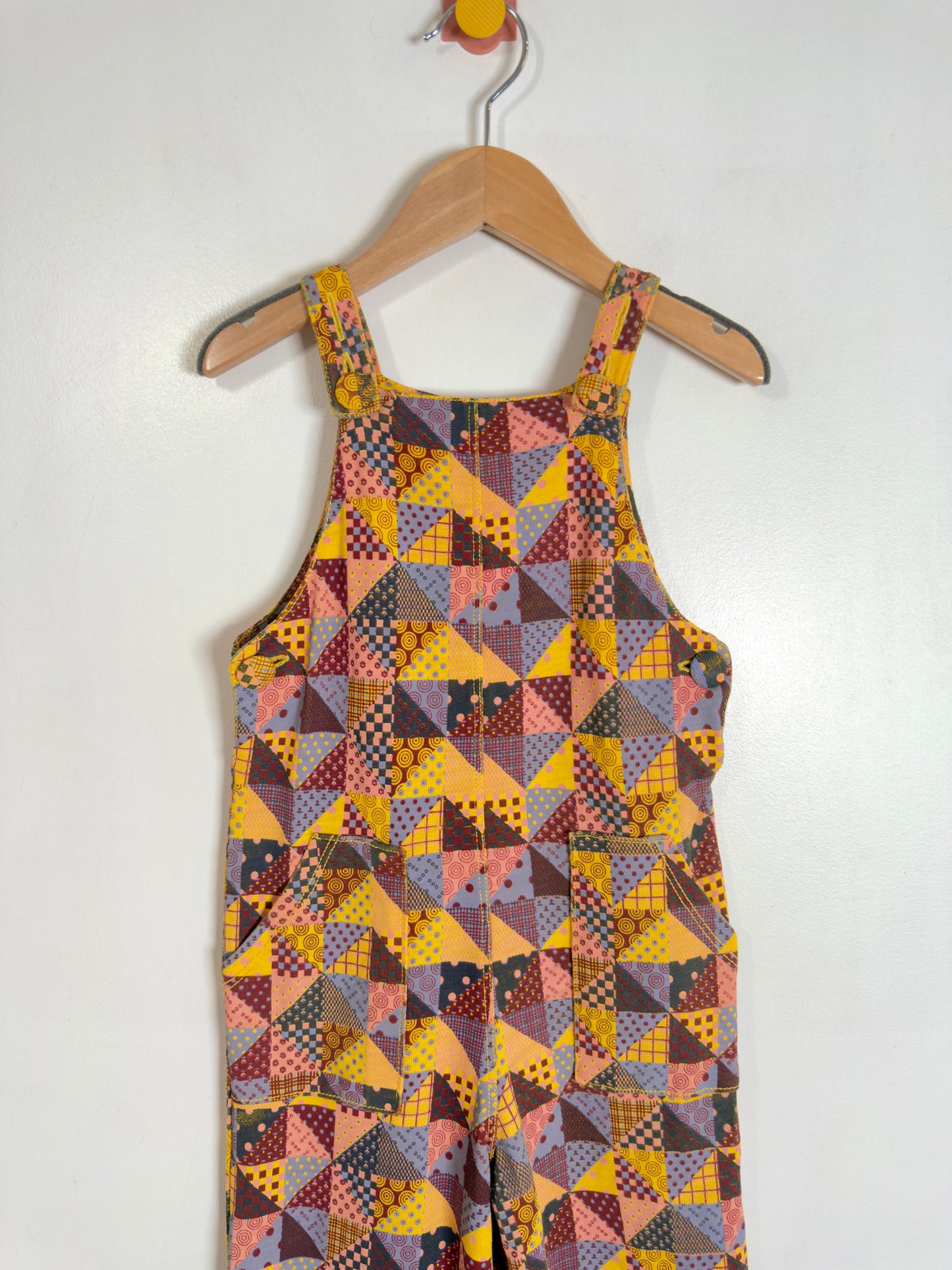 A sleeveless baby romper with a colorful geometric patchwork print, hanging on a wooden hanger against a plain white wall.