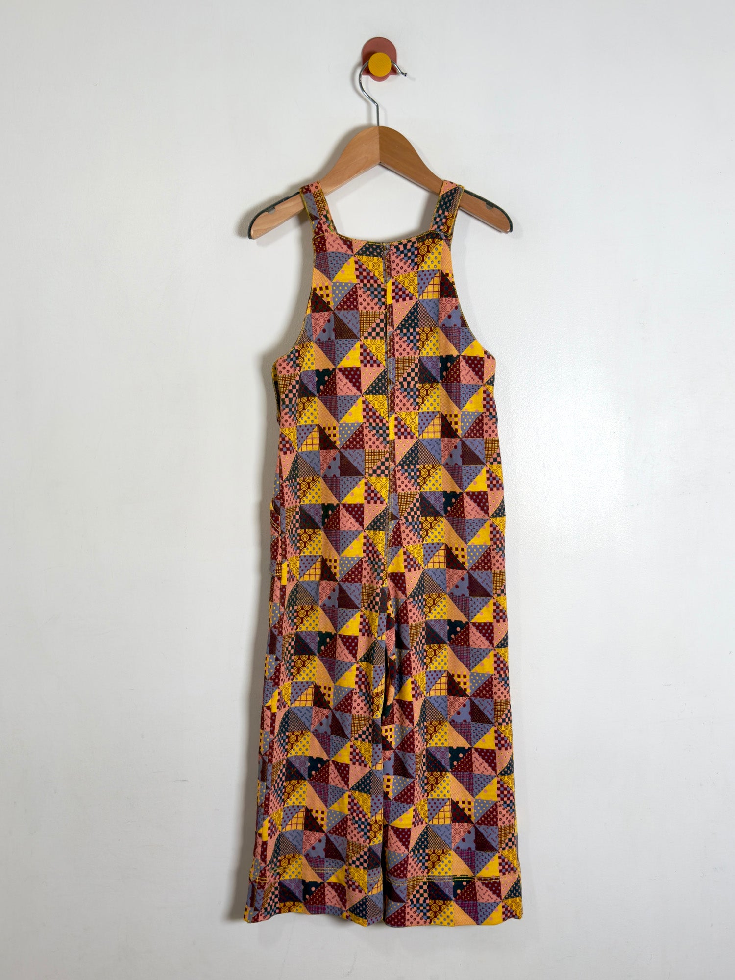 Overalls with a colorful geometric patchwork print, hanging on a wooden hanger against a plain white wall.
