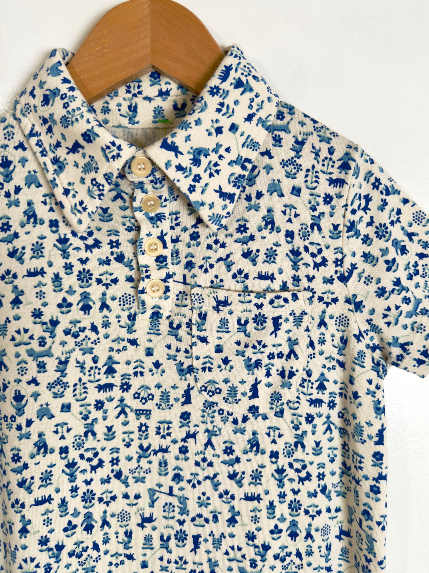 A short-sleeve button-up shirt with a blue and white hand-drawn style botanical pattern, hanging on a wooden hanger against a plain white background.