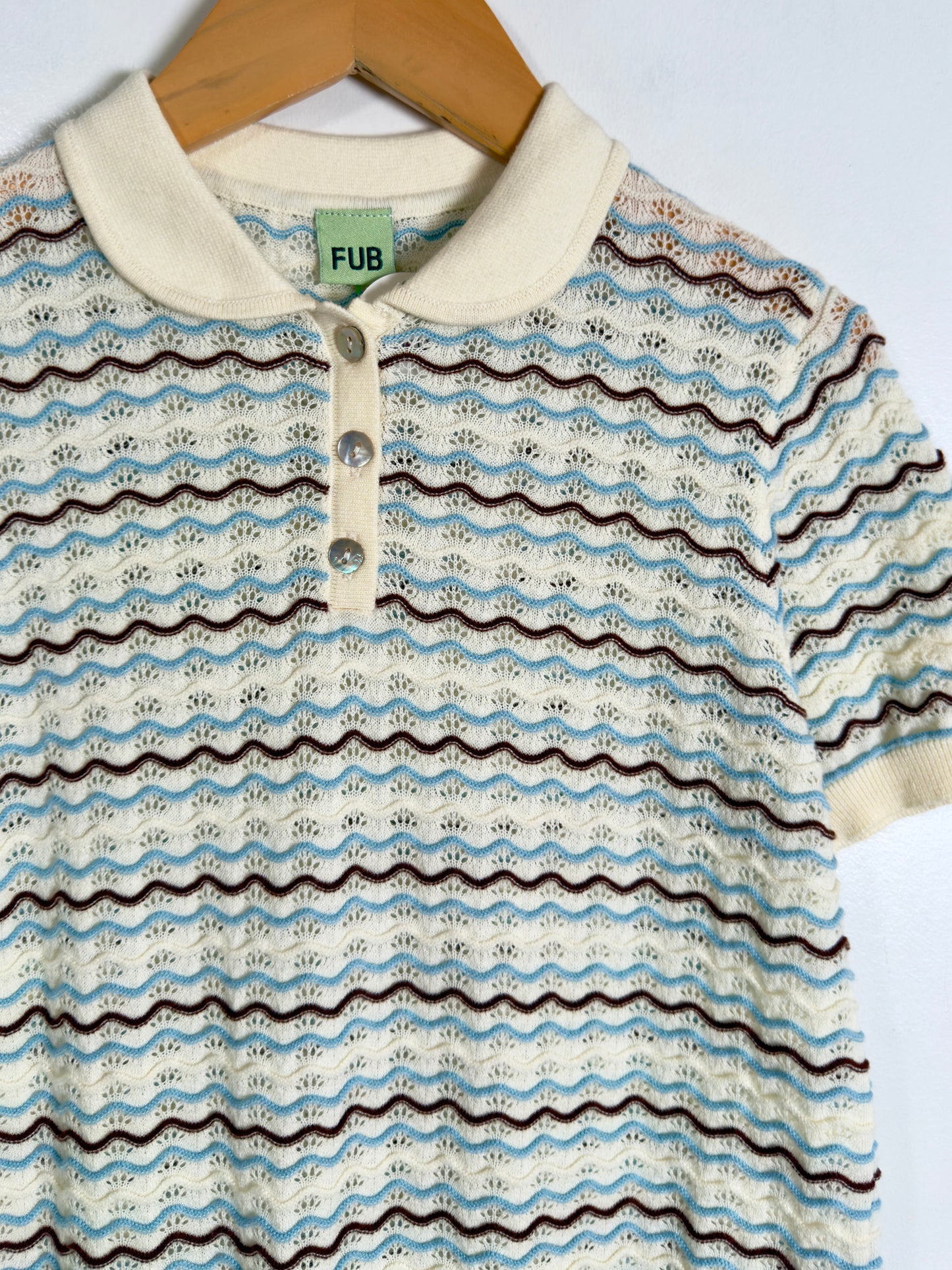 A short-sleeve knit polo shirt with a cream collar and three-button placket, featuring horizontal wavy stripes in light blue, dark brown, and black over an off-white base. The shirt is hanging on a wooden hanger against a plain white background, with a green FUB brand label visible inside the collar.