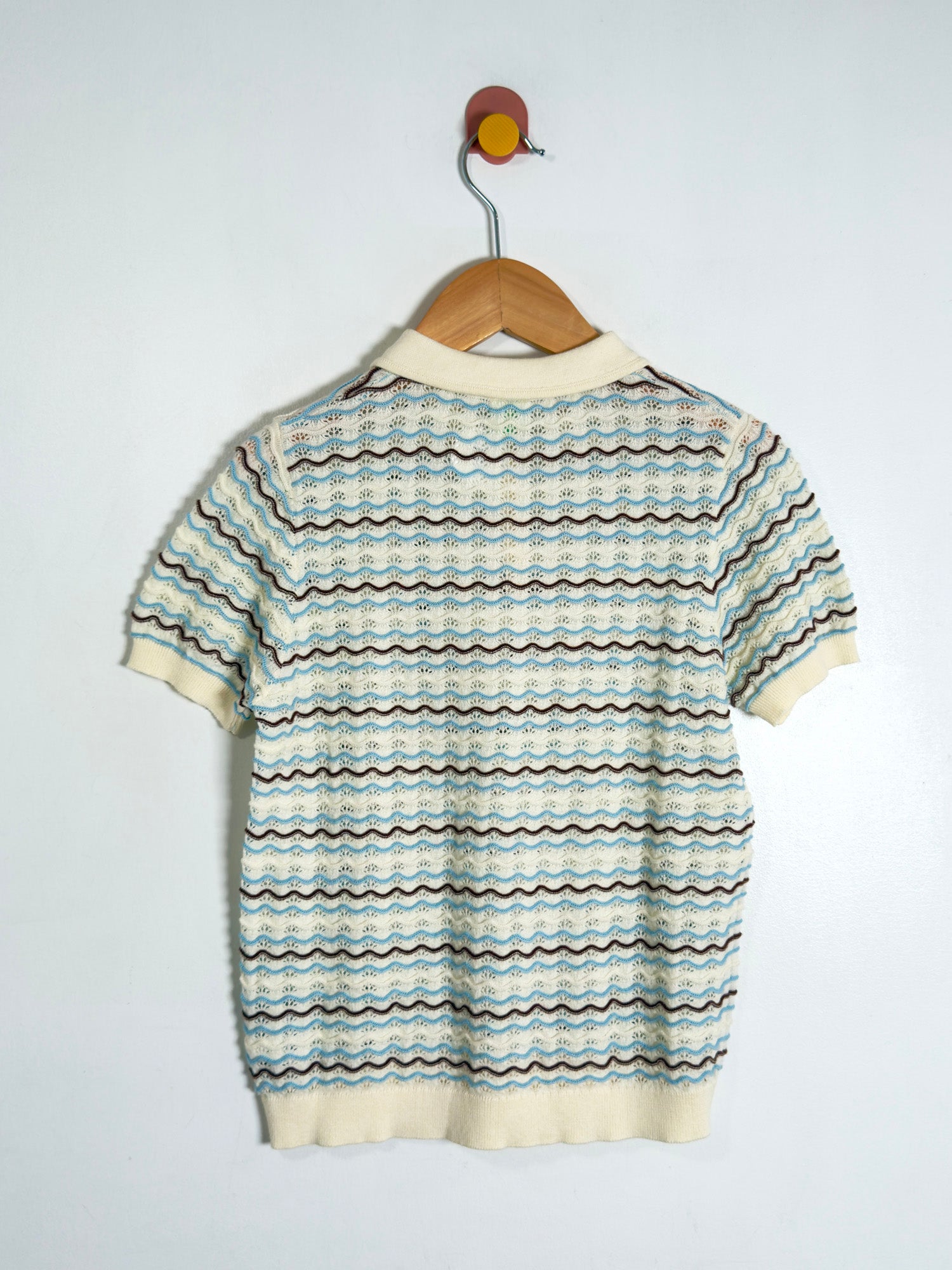 A short-sleeve knit polo shirt with a horizontal wavy stripe pattern in cream, light blue, and brown, hanging on a wooden hanger against a plain white wall.