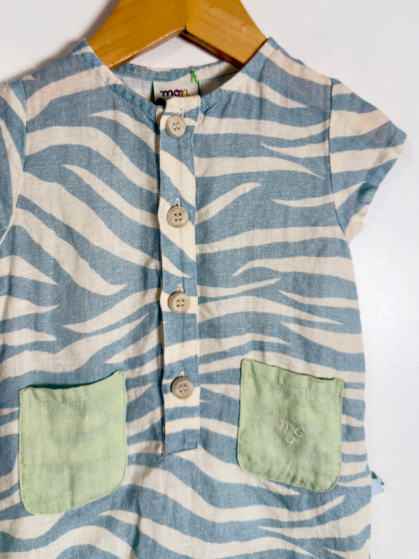 A children's short-sleeve top with a blue and off-white wavy stripe pattern, featuring a button-up front and two light green patch pockets, hanging on a wooden hanger against a white background.