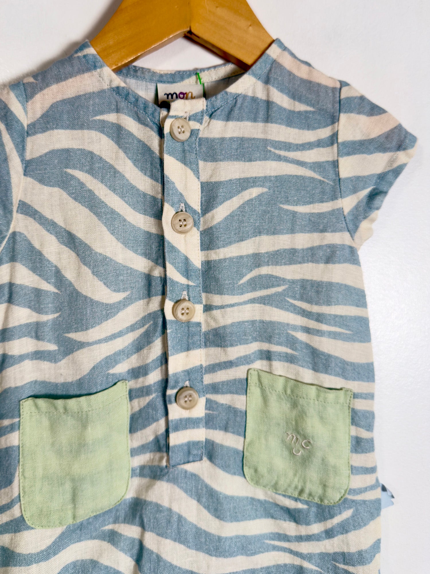 A children's short-sleeve top with a blue and off-white wavy stripe pattern, featuring a button-up front and two light green patch pockets, hanging on a wooden hanger against a white background.