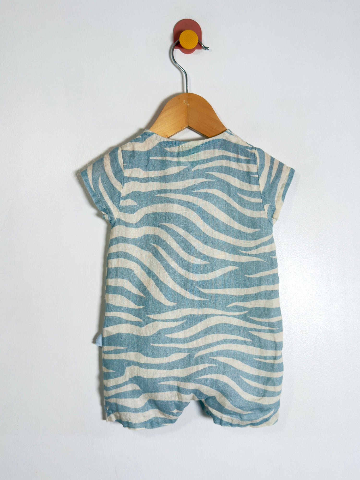 A baby short-sleeve romper with a wavy zebra-like pattern in dusty blue and cream, hanging on a wooden hanger against a plain white wall.