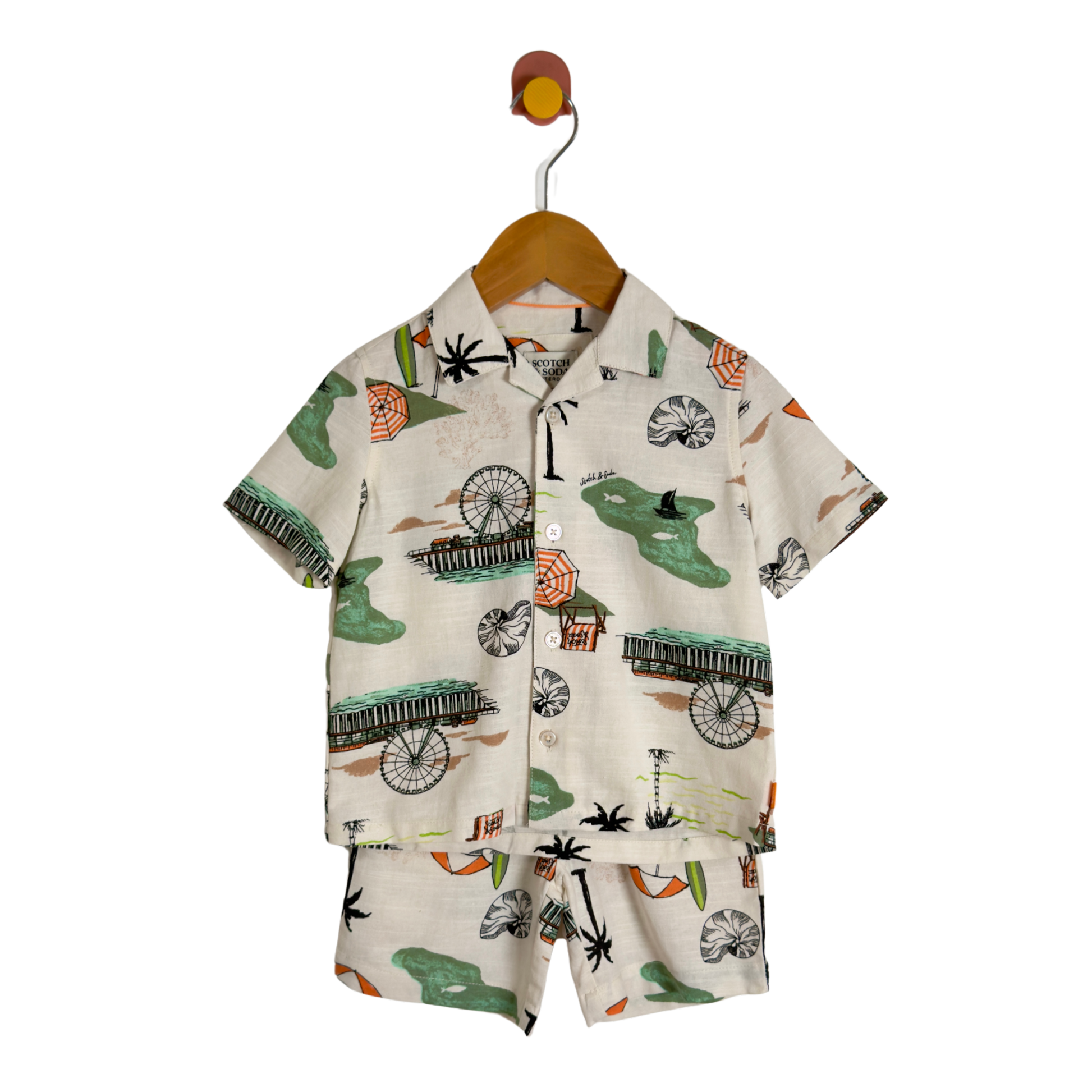 A two-piece boy's short-sleeve button-up shirt and matching shorts set, featuring a tropical beach print with palm trees, beach umbrellas, and green palm trees on a cream-colored background, hanging on a wooden hanger.