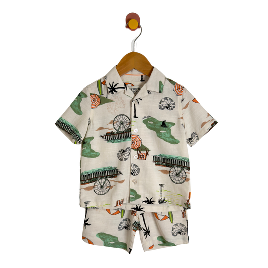 A two-piece boy's short-sleeve button-up shirt and matching shorts set, featuring a tropical beach print with palm trees, beach umbrellas, and green palm trees on a cream-colored background, hanging on a wooden hanger.