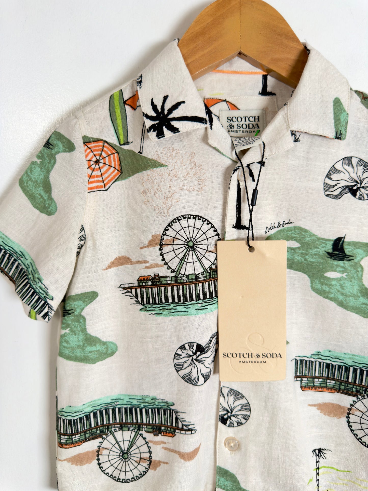 A short-sleeve button-up shirt by Scotch & Sofia hanging on a wooden hanger against a white wall. The shirt has an off-white base with a tropical print featuring green palm trees and waves, black Ferris wheels and umbrellas, and orange beach umbrellas.