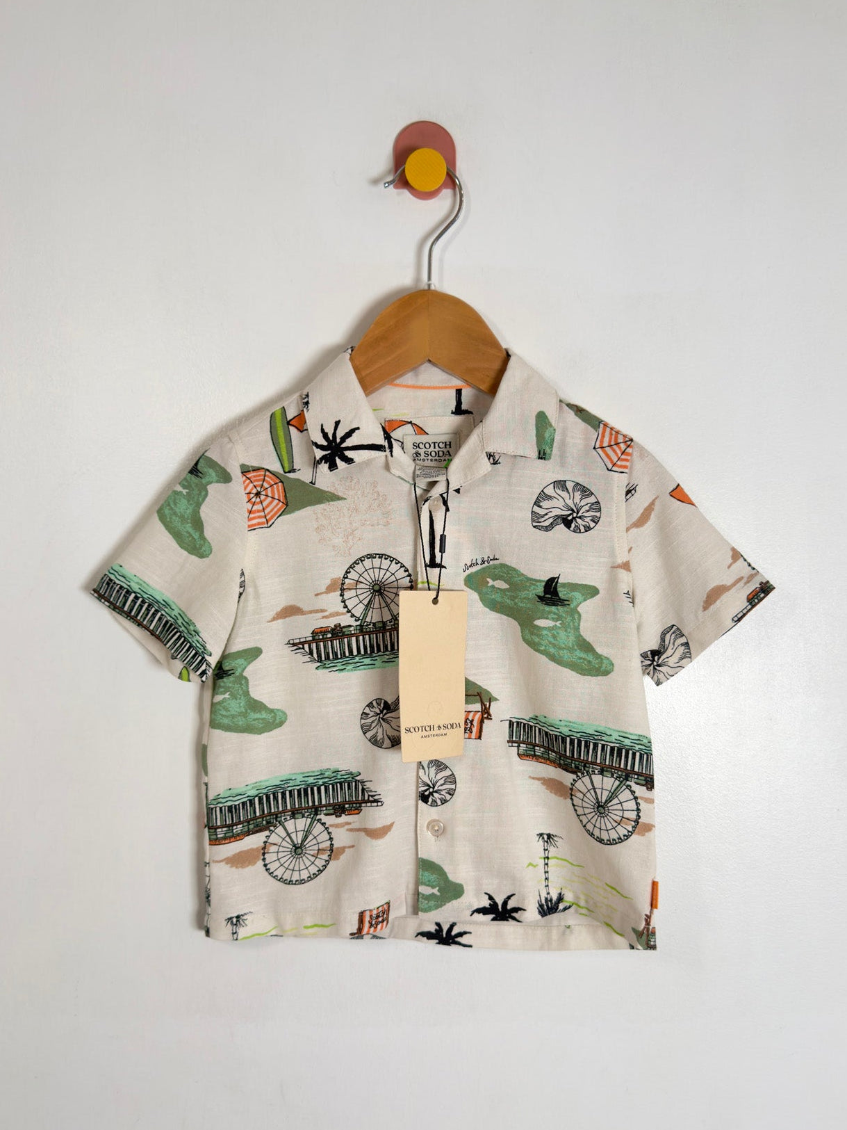A short-sleeve button-up shirt for kids, featuring a print  on a cream-colored background. The shirt is displayed on a wooden hanger with a paper tag, hanging from a wall hook.