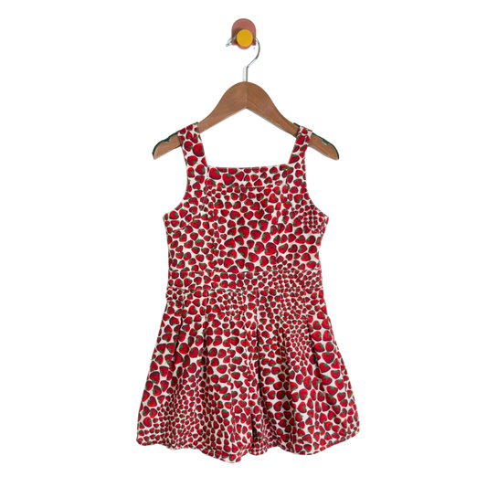A sleeveless children's dress with a red, white, and black spotted pattern, hanging on a wooden hanger against a plain white background.