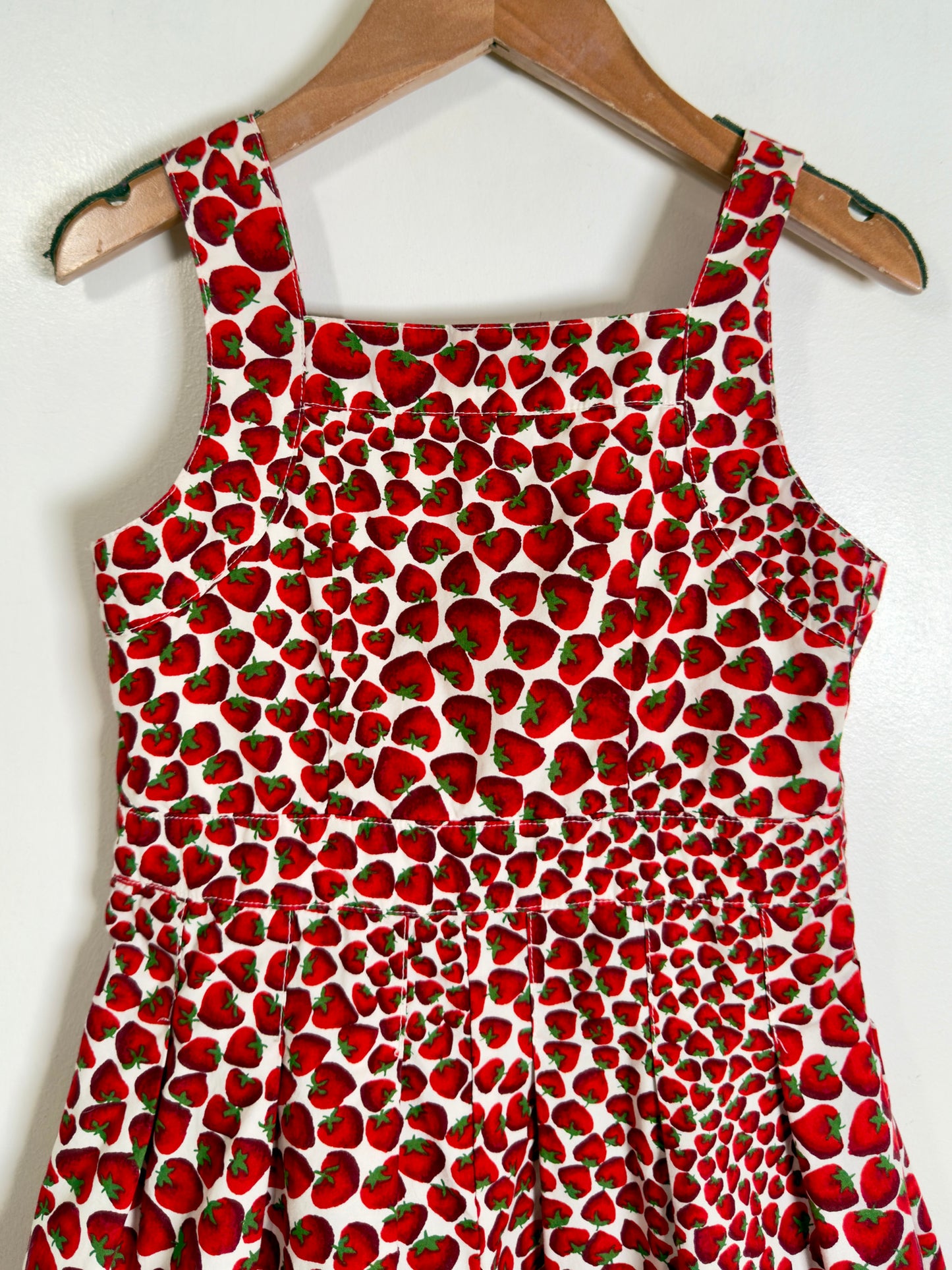 A sleeveless white dress with an all-over red strawberry print, hanging on a wooden hanger against a plain white wall.