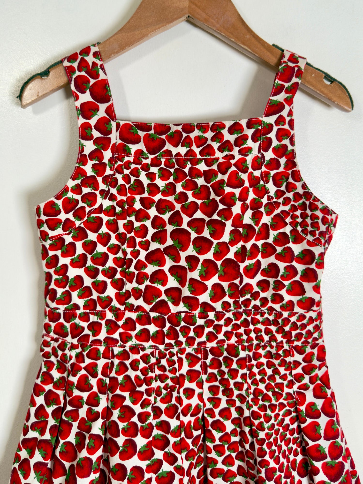 A sleeveless white dress with an all-over red strawberry print, hanging on a wooden hanger against a plain white wall.