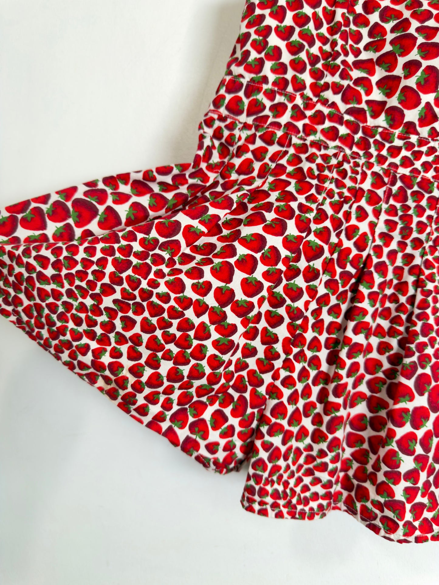 A close-up of a white fabric garment, likely a skirt or shorts, featuring an all-over red strawberry print with green leaves on a plain white background.