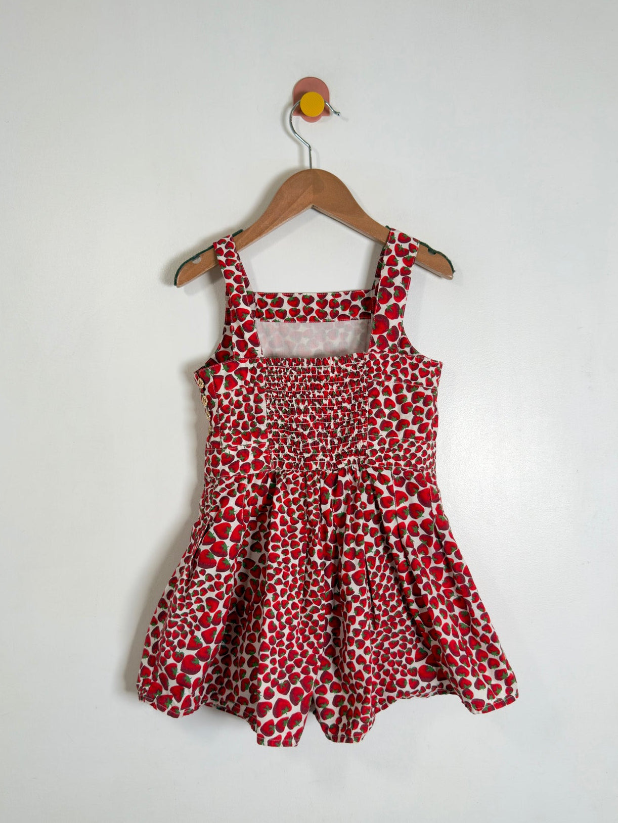 A sleeveless red and white patterned dress with a smocked bodice and flared skirt, hanging on a wooden hanger against a plain white wall.