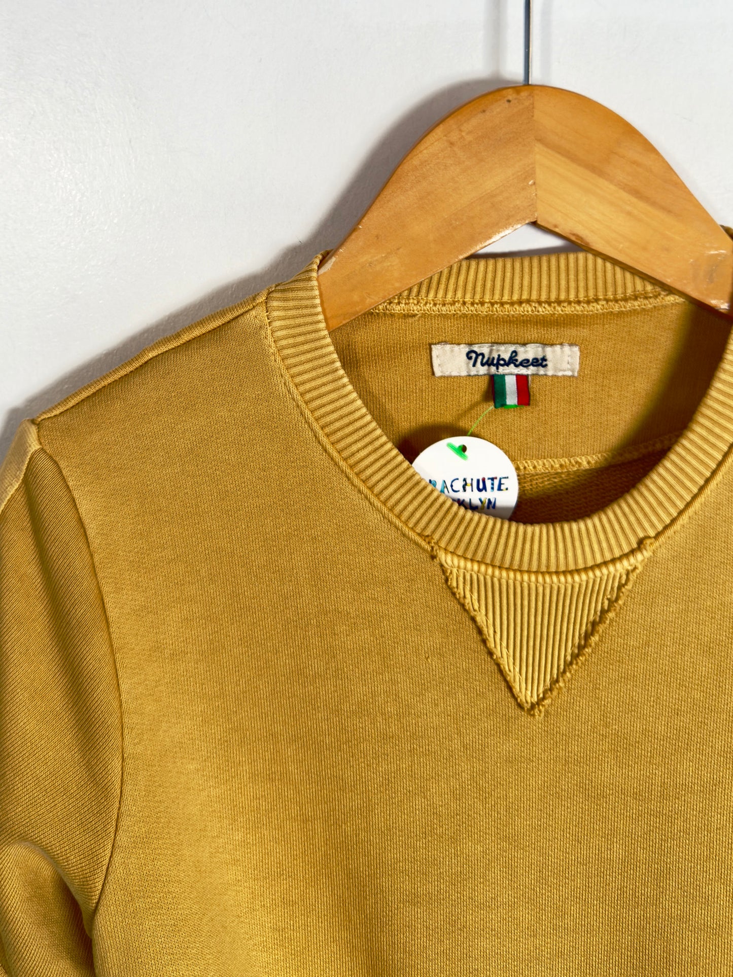 A mustard yellow ribbed knit sweater hanging on a wooden hanger against a plain light gray wall. The neck label reads 'Pisshouse' and a hangtag is attached.