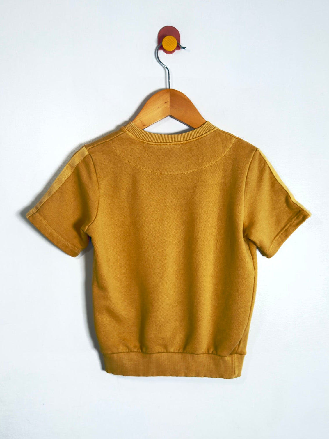 A mustard yellow short-sleeve crewneck sweatshirt hanging on a wooden hanger against a plain white wall.