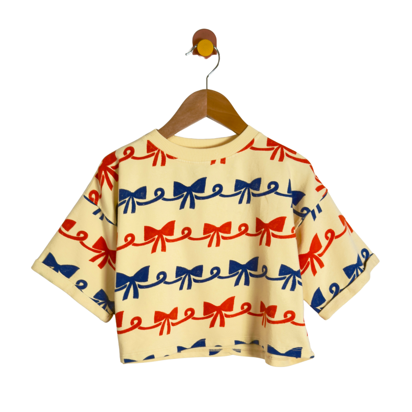 A short-sleeve baby tee with a repeating red and blue bow pattern on a cream-colored fabric, hanging on a wooden hanger against a solid white background.
