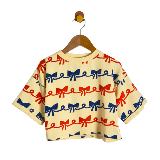 A short-sleeve baby tee with a repeating red and blue bow pattern on a cream-colored fabric, hanging on a wooden hanger against a solid white background.