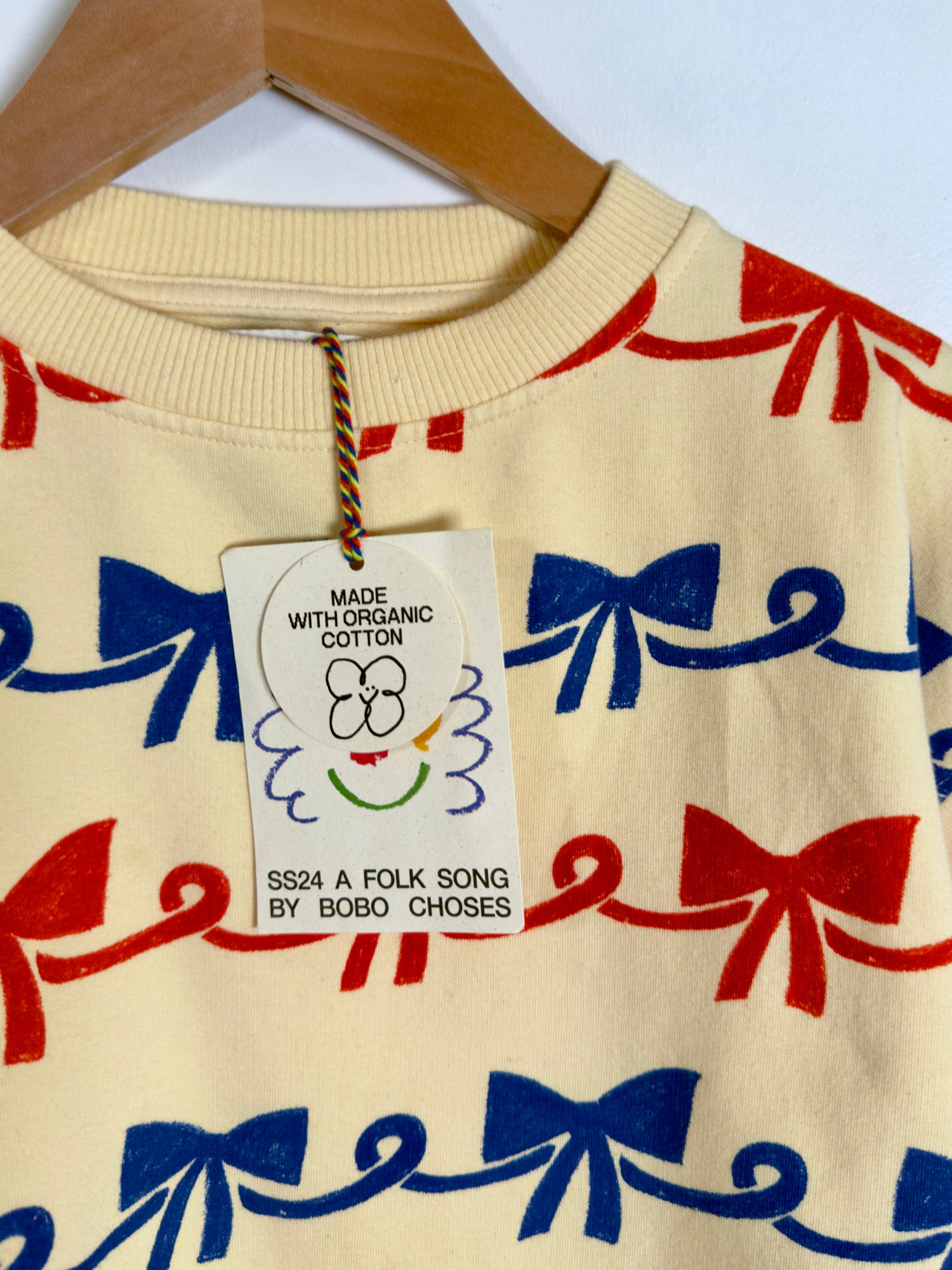 A cream-colored crewneck sweatshirt with a repeating pattern of red and blue bows and scroll motifs, hanging on a wooden hanger with its original product tag attached.