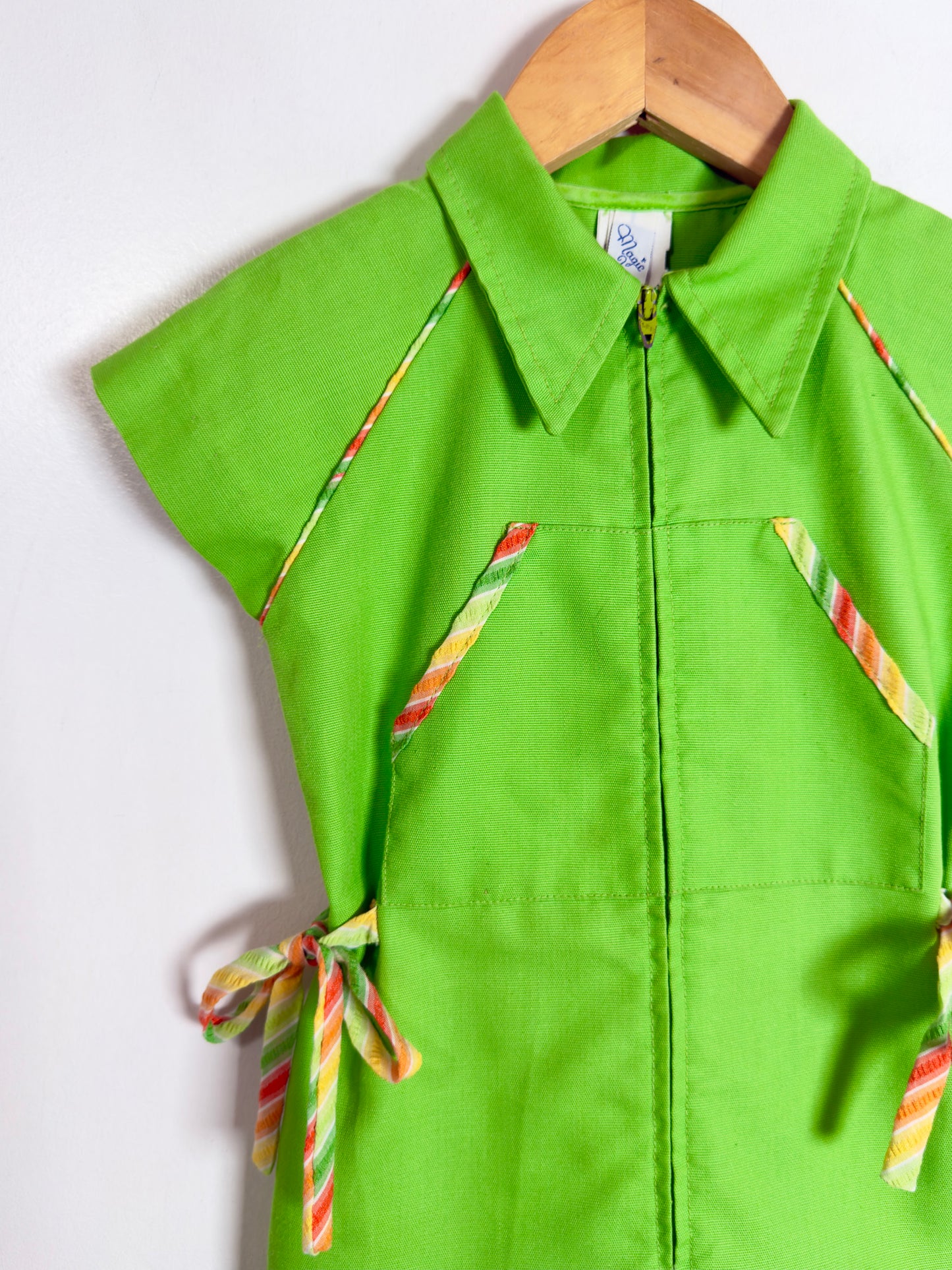 A bright lime green short-sleeve collared shirt dress with a front zipper, featuring diagonal striped tie details in orange, yellow, and green on the chest and sides, hanging on a wooden hanger against a white wall.