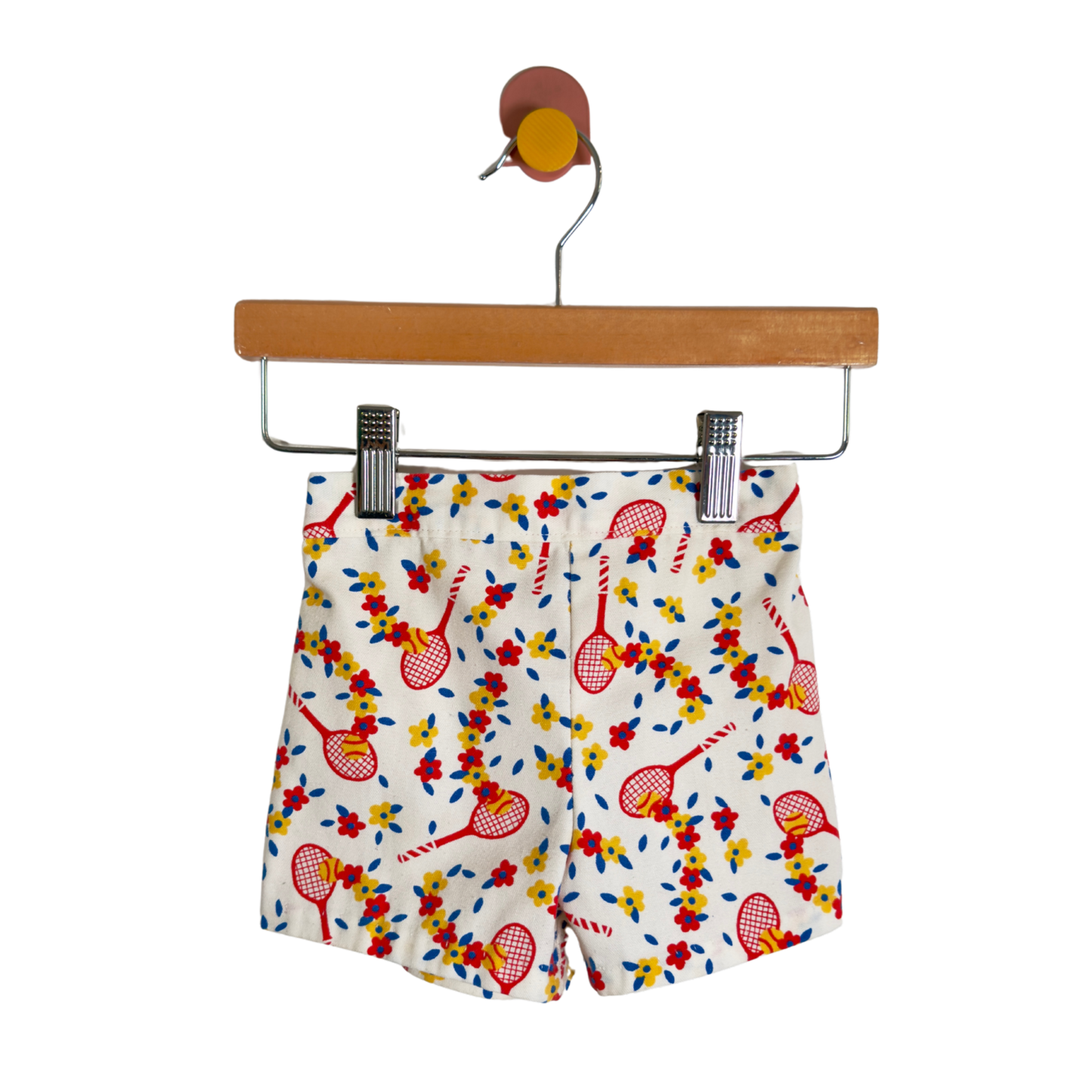 White shorts with a colorful tennis racket and floral pattern, hanging from a wooden hanger against a white background.