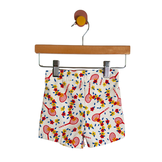White shorts with a colorful tennis racket and floral pattern, hanging from a wooden hanger against a white background.