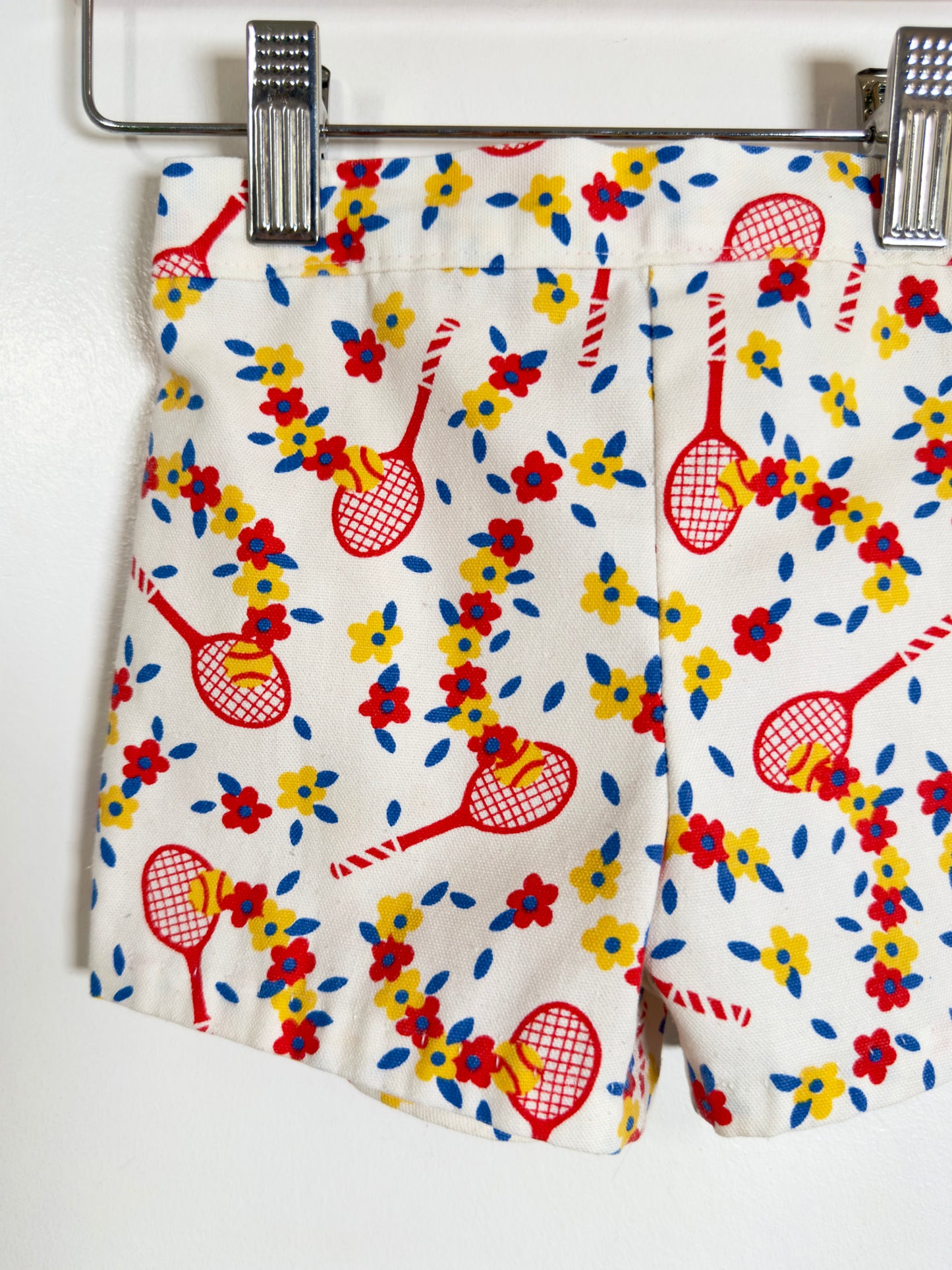 White shorts with a colorful tennis racket and floral print, hanging from a metal hanger clip against a white wall.