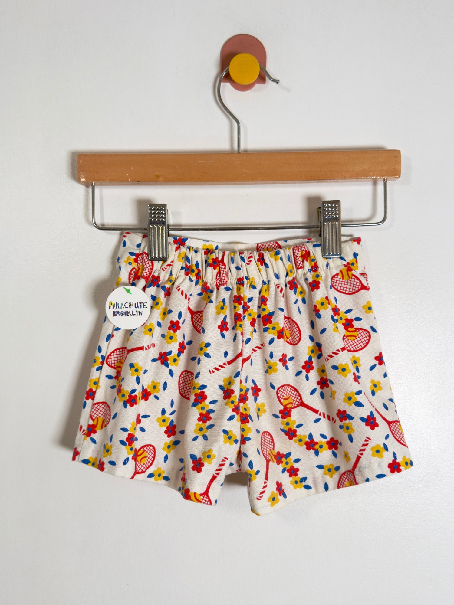 White shorts with a colorful tennis racket and floral print, hanging from a wooden hanger with metal clips against a plain white wall.