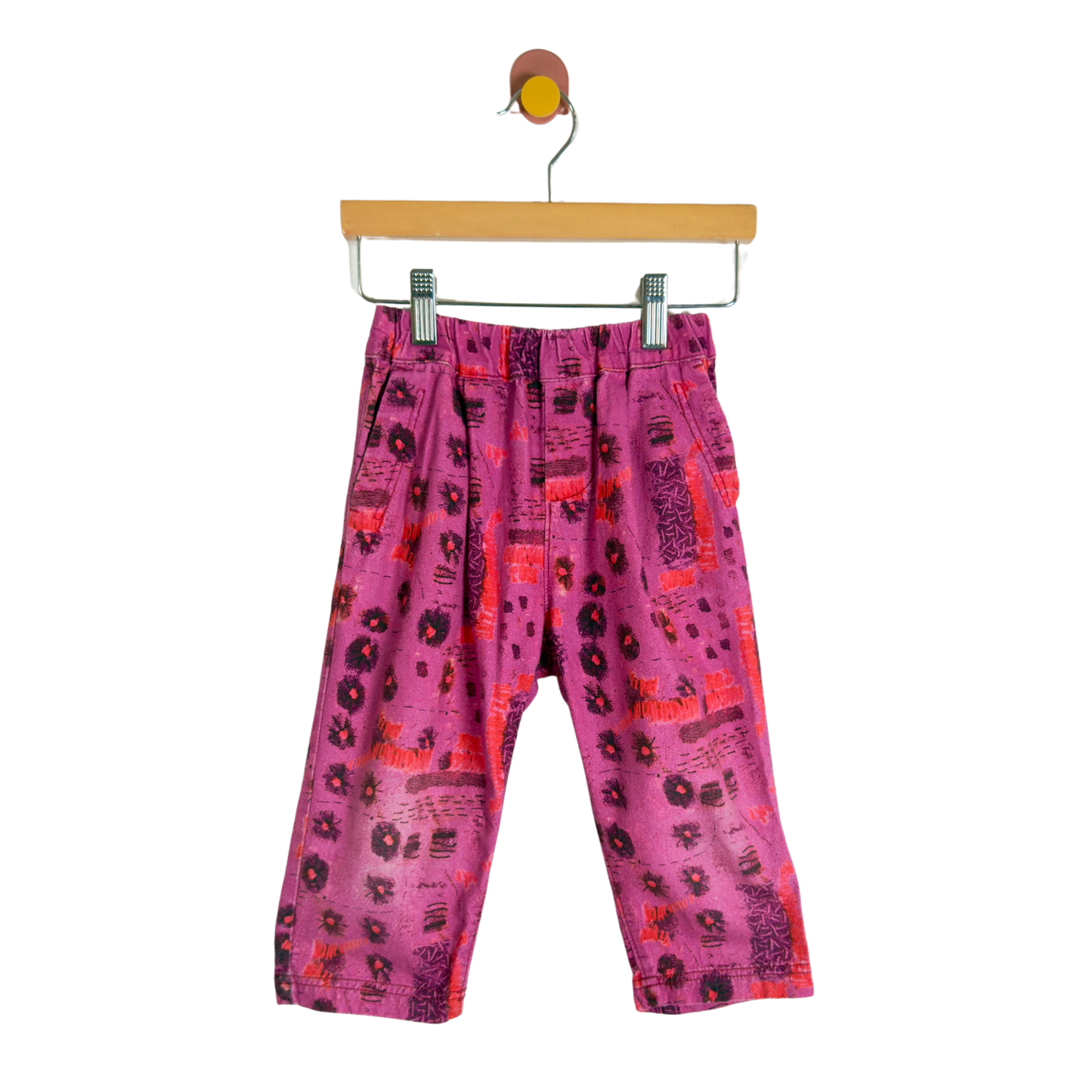 Pink patterned children's pants with black and red abstract prints, hanging from a wooden hanger against a white background.