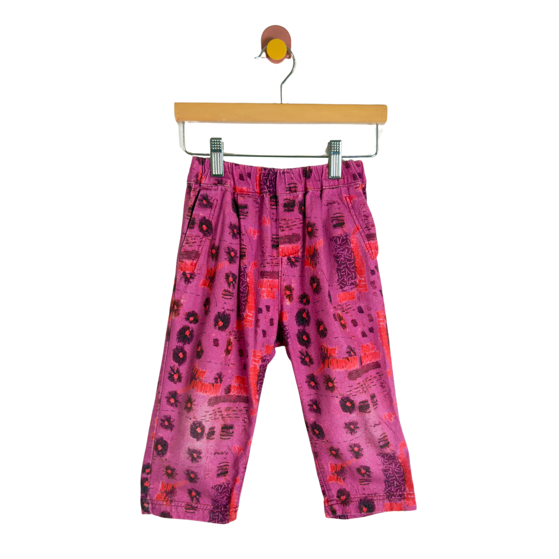 Pink patterned children's pants with black and red abstract prints, hanging from a wooden hanger against a white background.