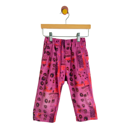 Pink patterned children's pants with black and red abstract prints, hanging from a wooden hanger against a white background.