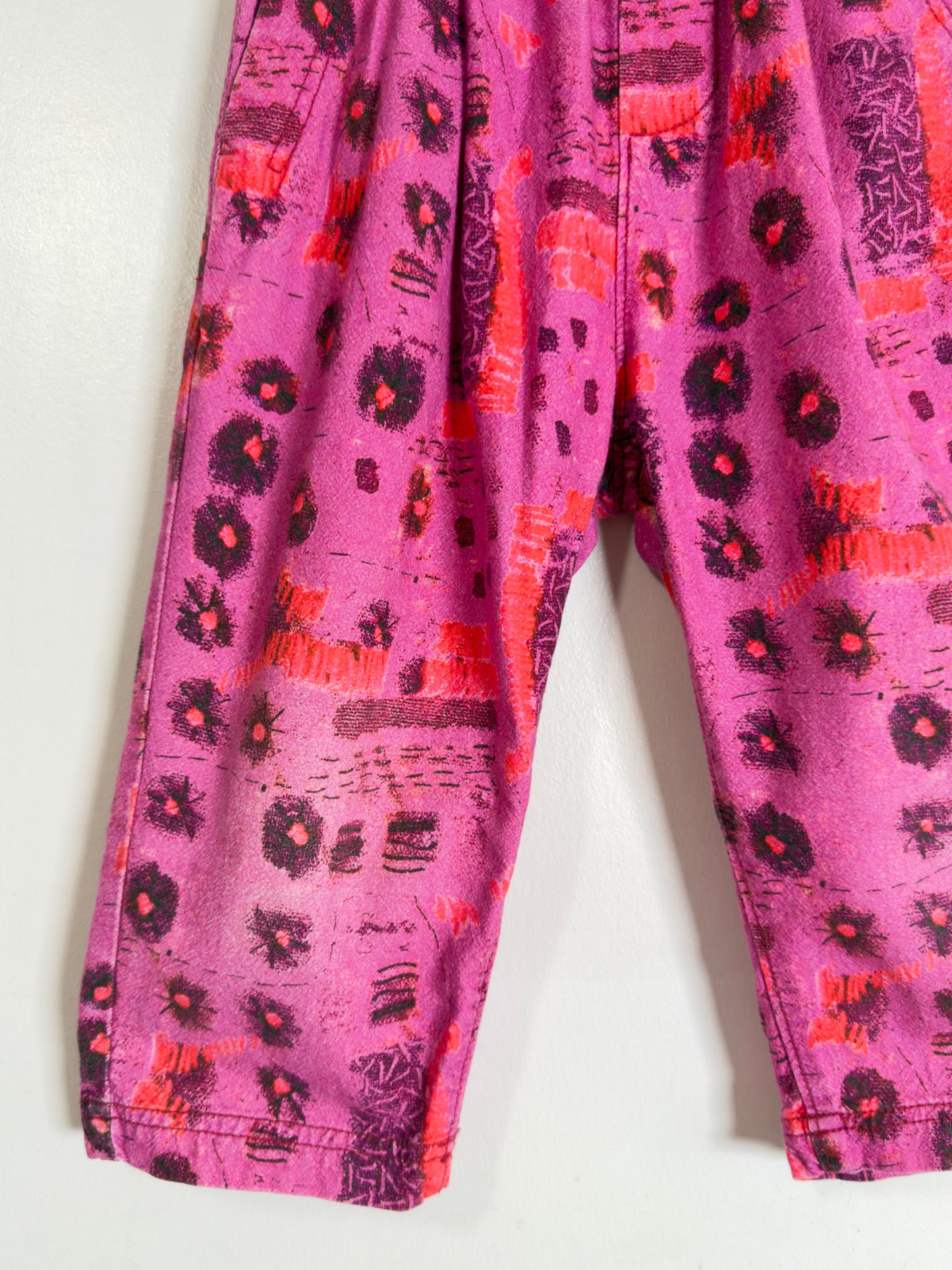 Pink patterned pants with orange and black abstract floral and brushstroke designs, shown hanging against a plain light background.