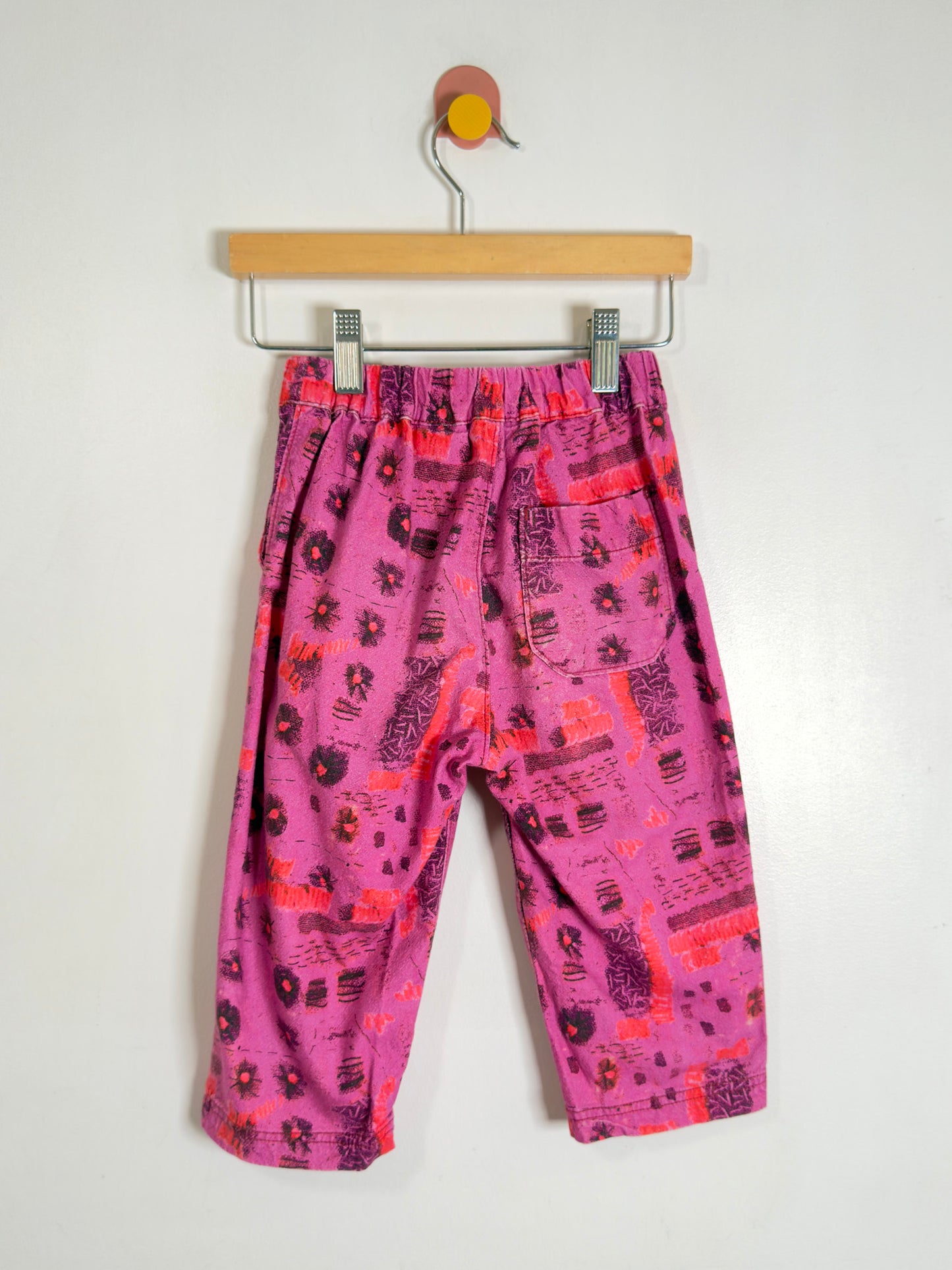 Pink patterned pants with orange and black abstract prints, hanging from a wooden hanger against a plain white wall.