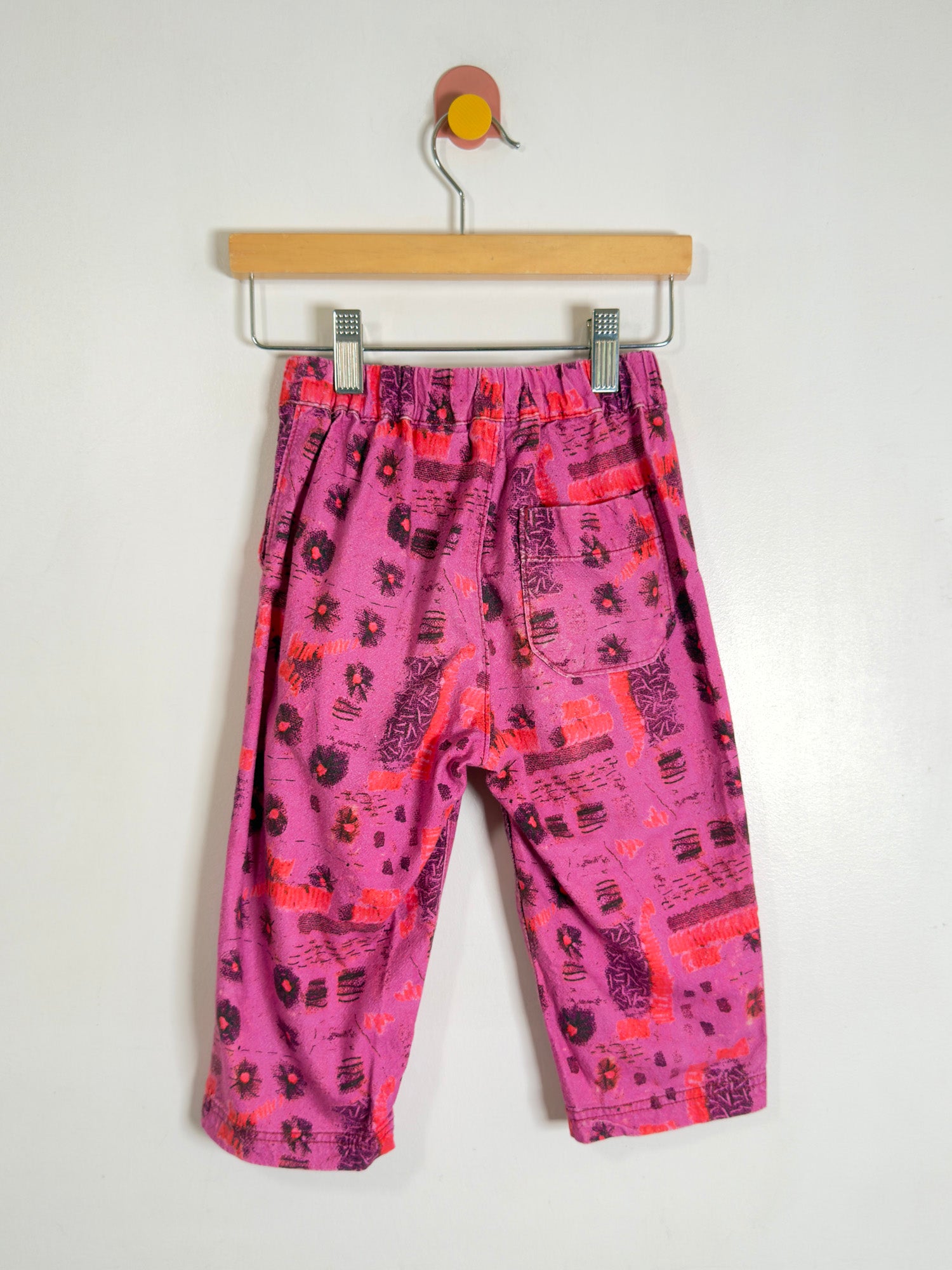 Pink patterned pants with orange and black abstract prints, hanging from a wooden hanger against a plain white wall.