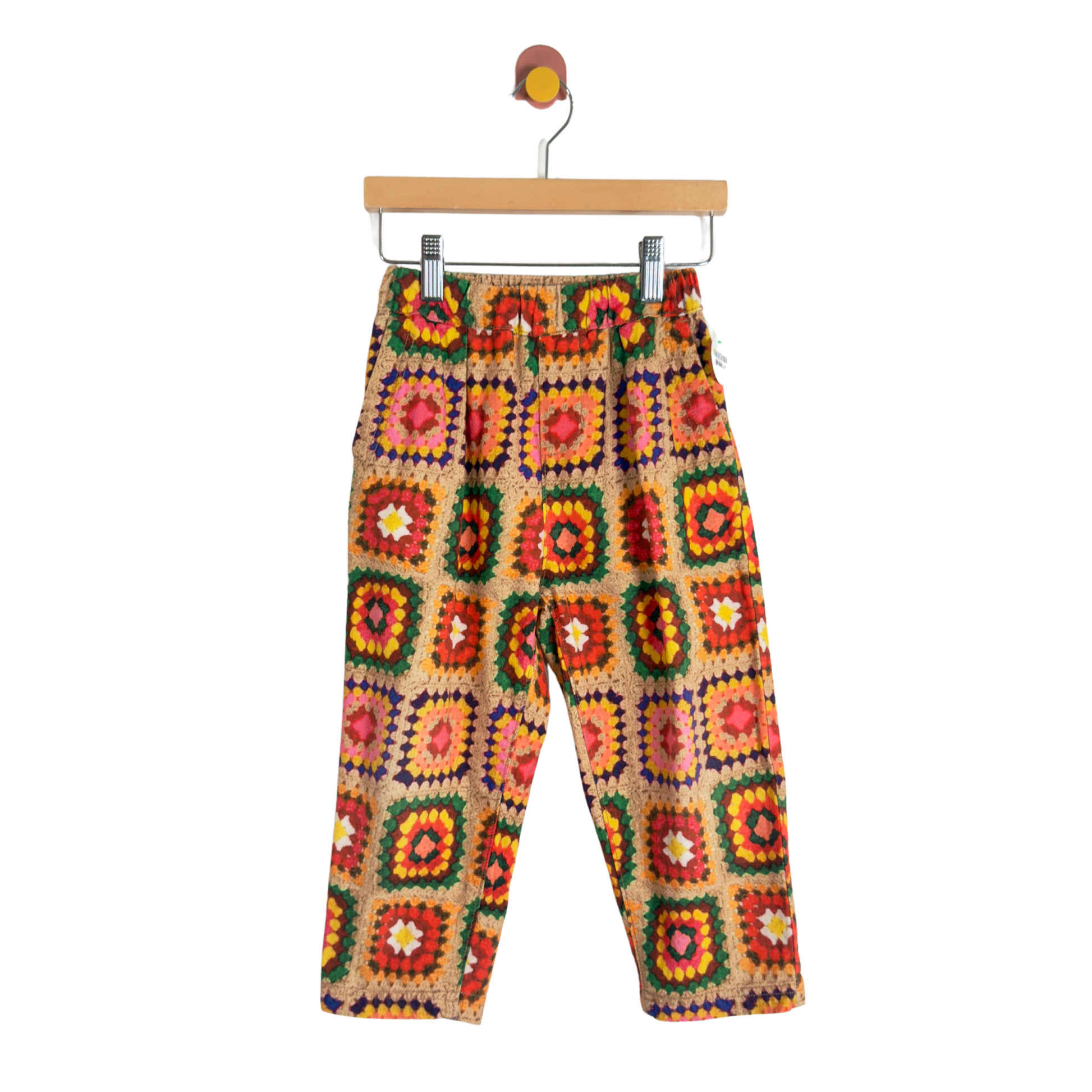 A pair of children's pants with a colorful crochet granny-square pattern, hanging from a wooden hanger against a plain white background.