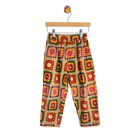 A pair of children's pants with a colorful crochet granny-square pattern, hanging from a wooden hanger against a plain white background.