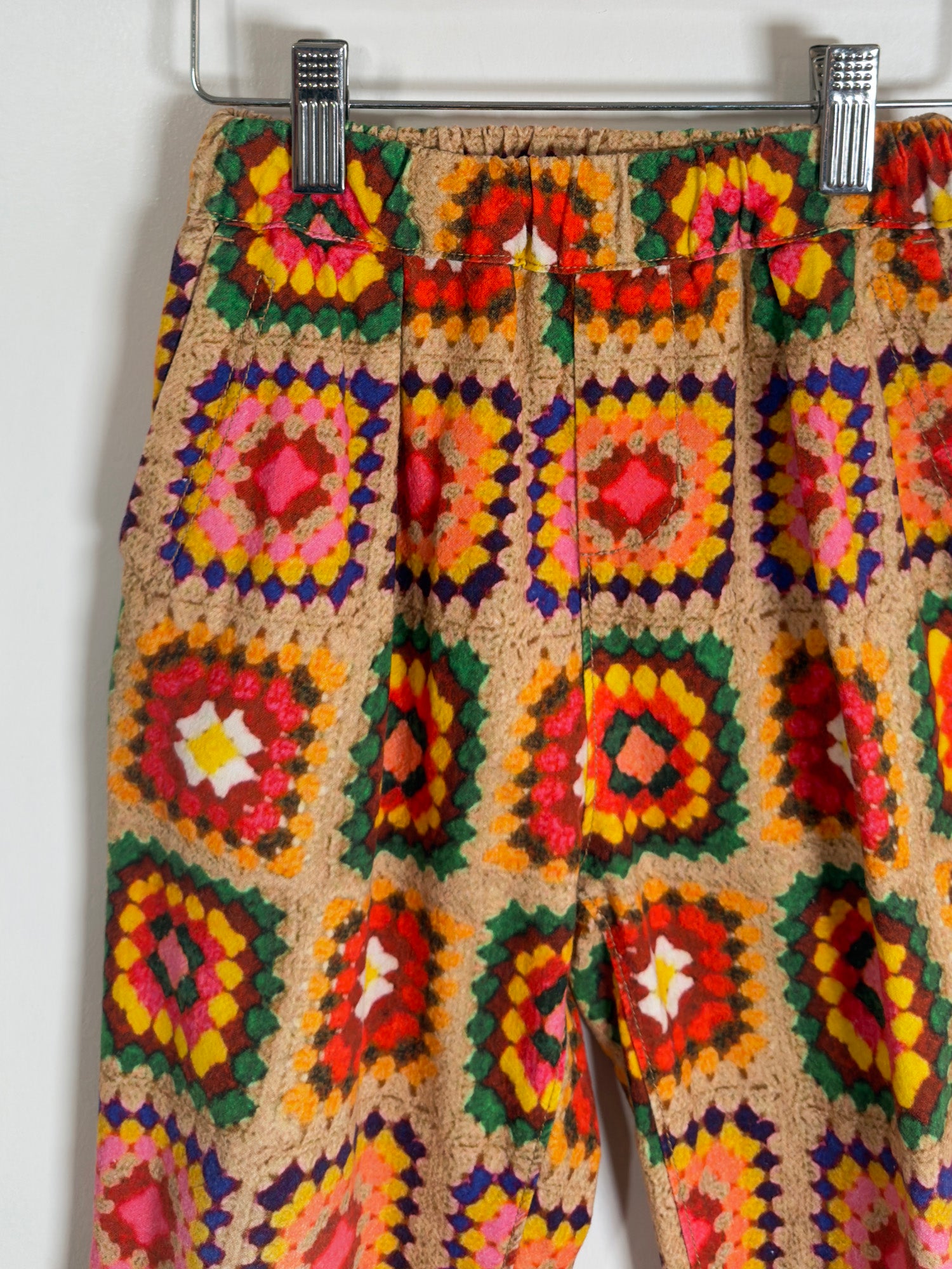 A pair of cropped pants featuring a colorful crochet granny square pattern on a beige base, hanging from a metal rack with two silver clips against a plain white wall.