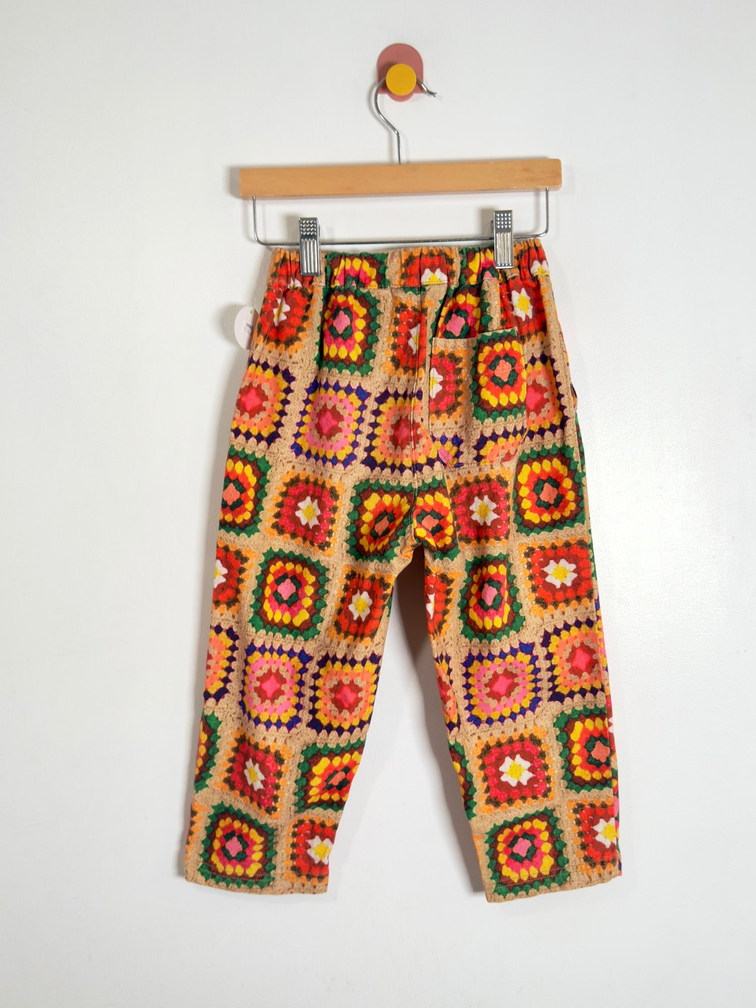 A pair of children's pants with a colorful crocheted granny-square pattern on a beige base, hanging from a wooden hanger against a plain white wall.