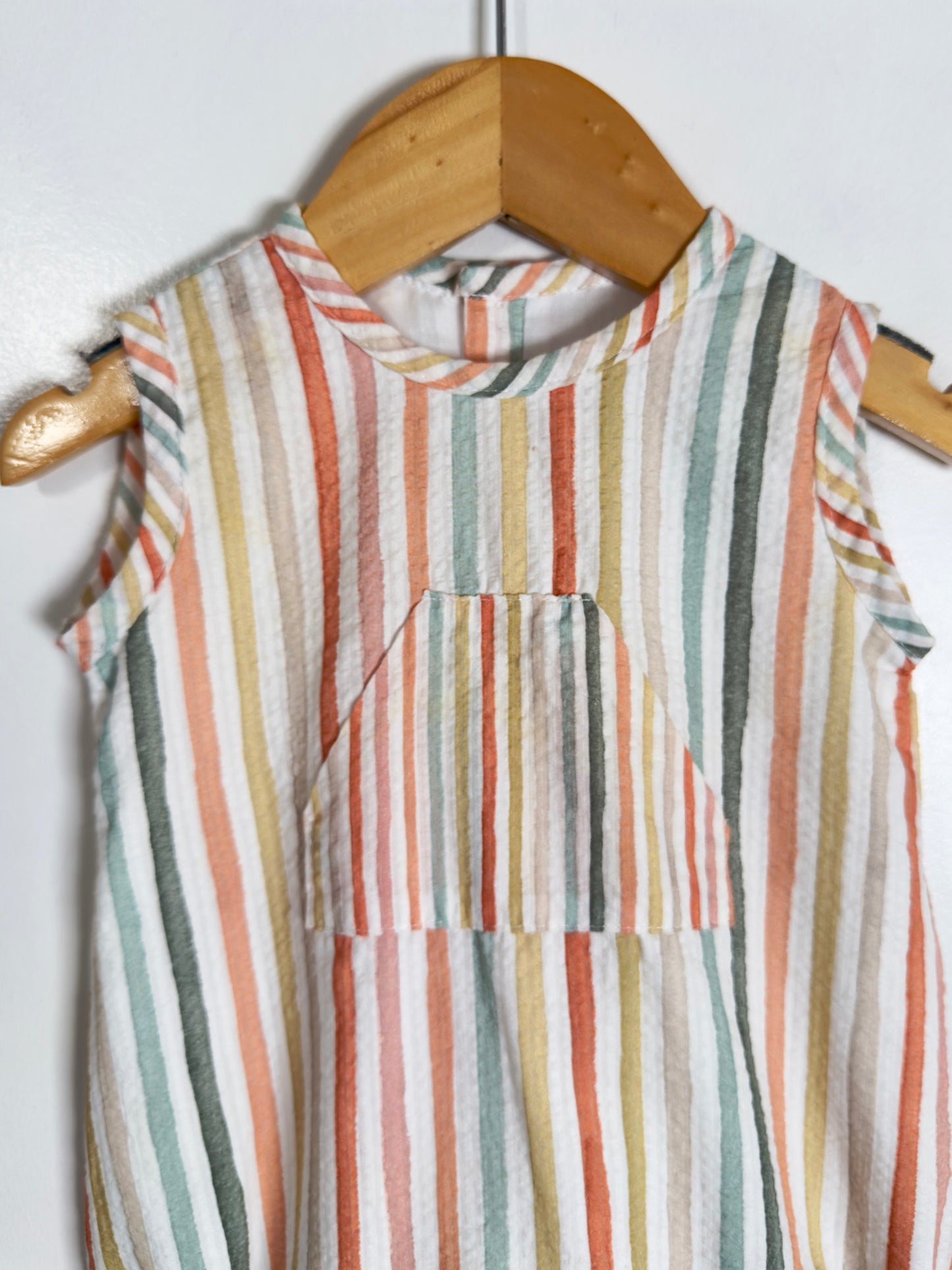 A sleeveless top with vertical stripes in muted orange, sage green, beige, and white, hanging on a wooden hanger against a plain white wall.